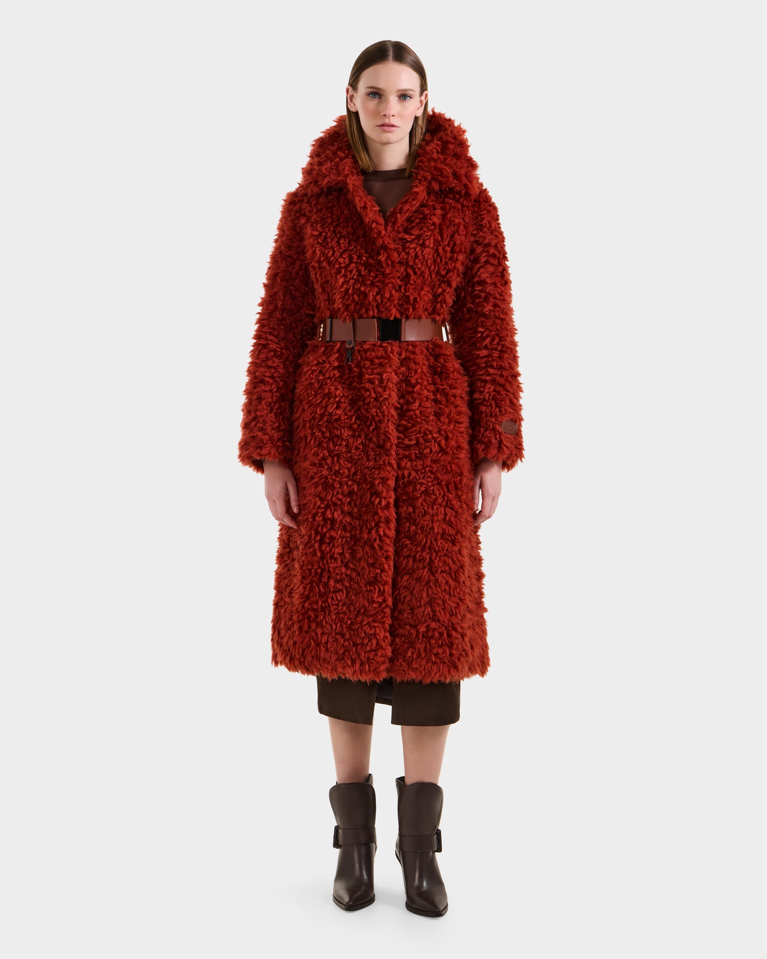 A woman stands facing forward against a plain white background wearing the KAMILAH - 8125609 Rust faux fur trench by Rudsak, styled with a vegan leather belt, brown mid-calf boots, and a brown dress underneath.