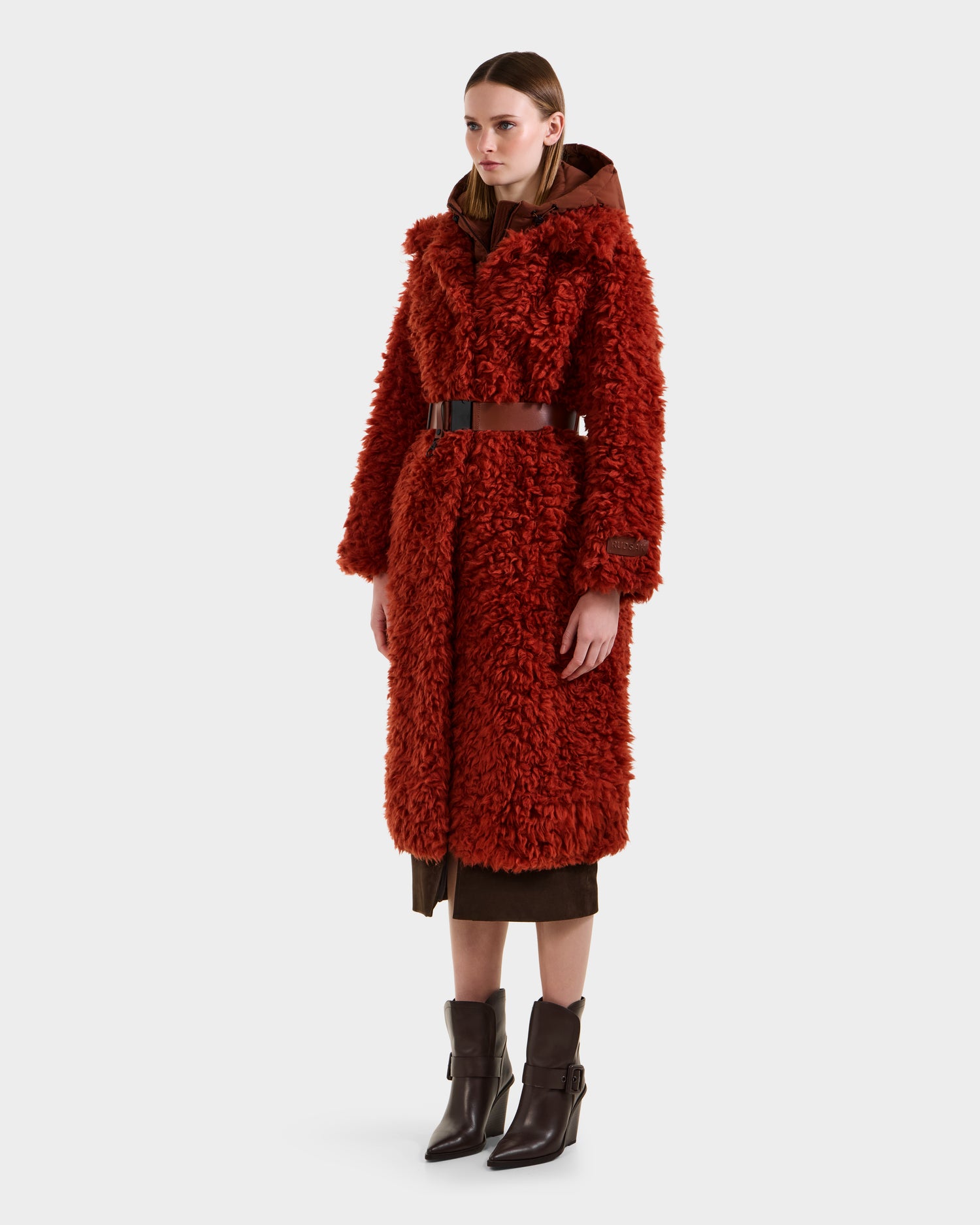 A woman wears the KAMILAH - 8125609 Rust faux fur trench by Rudsak, featuring a vegan leather belt and layered brown clothing with pointed brown ankle boots, set against a plain white background.