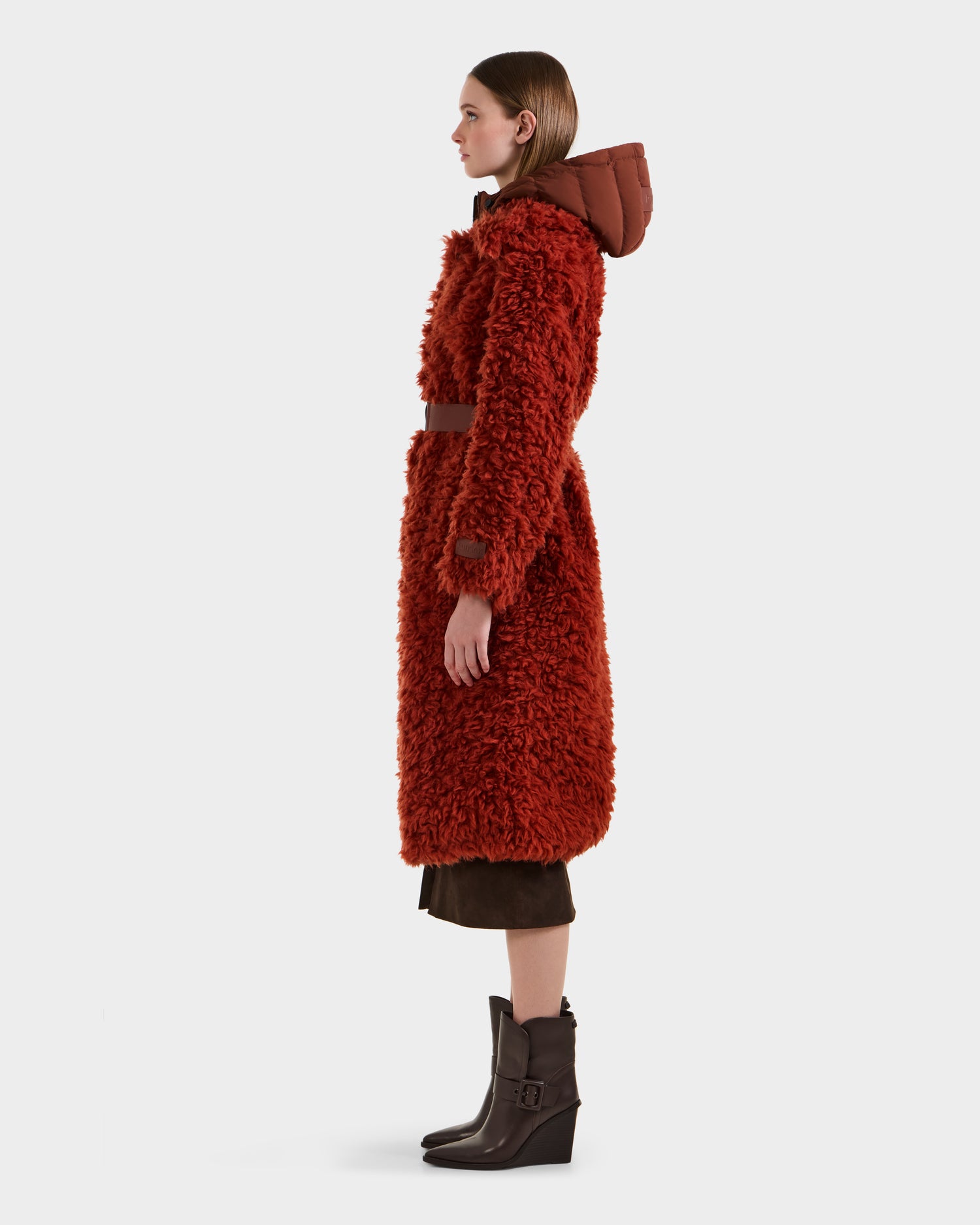 A woman stands in profile against a plain white background wearing the Rudsak KAMILAH - 8125609 Rust faux fur trench with a hood, styled with dark brown pants and ankle boots. The coat features a luxurious wool teddy texture.
