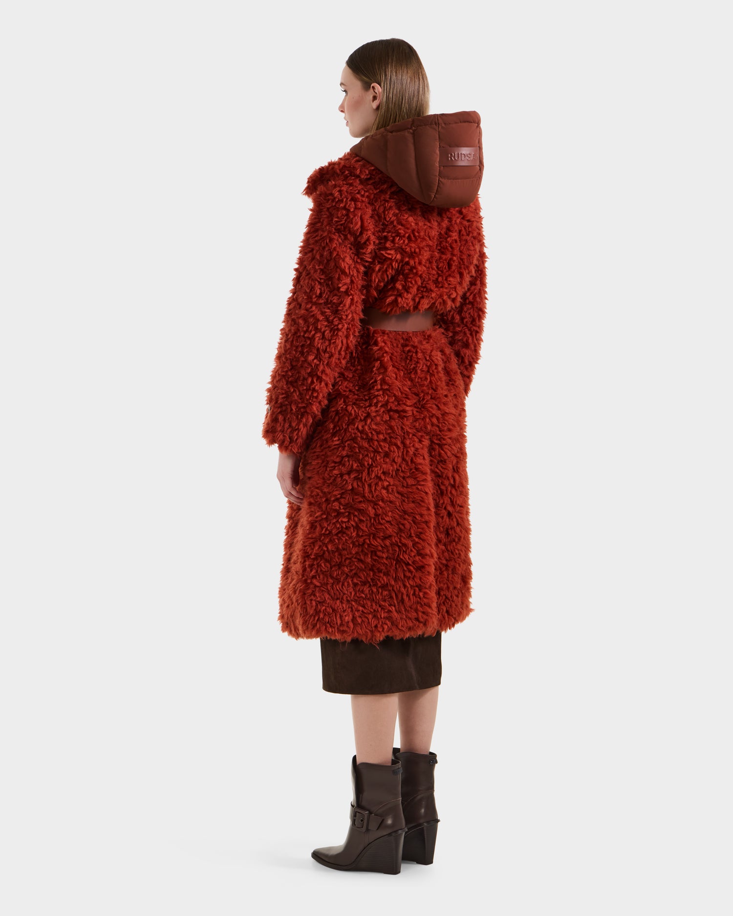 A person in the Rudsak KAMILAH - 8125609 Rust faux fur trench with a brown hood, straight brown hair, a brown skirt, and heeled boots stands facing away against a plain background.