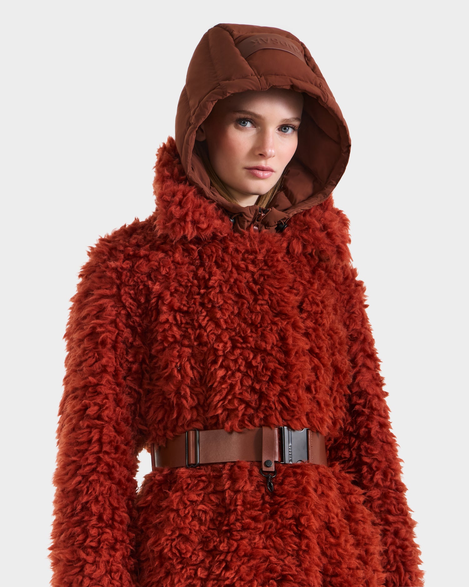 A woman wearing the KAMILAH - 8125609 Rust faux fur trench by Rudsak, featuring a vegan leather belt and brown hood, stands against a plain white background.