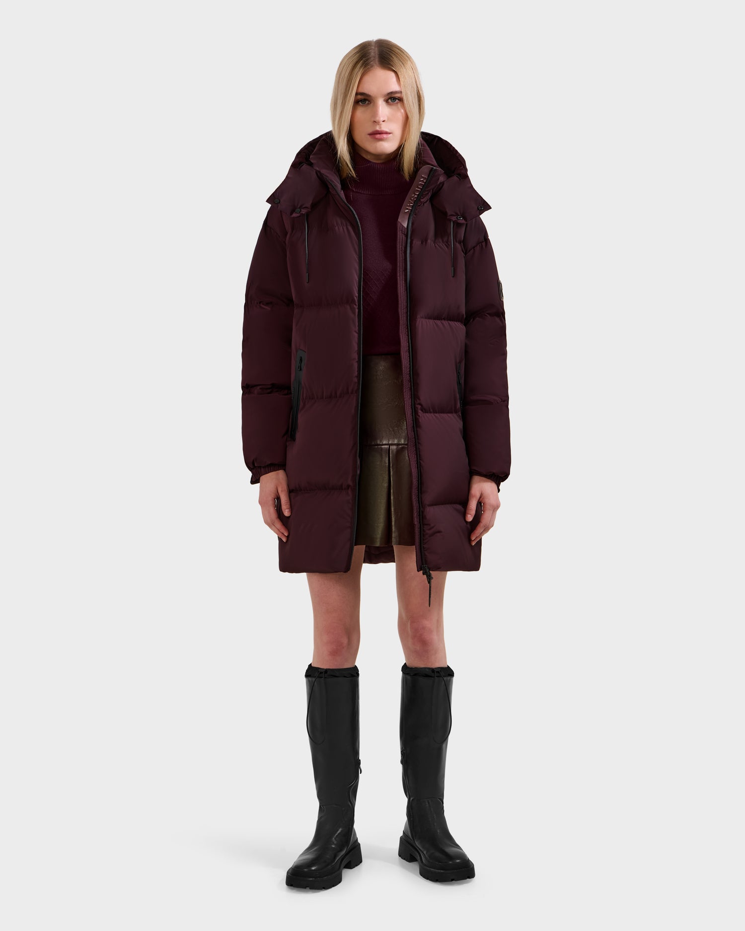 A woman stands facing forward in a Rudsak KARLEE L - 8125682 Bordeaux long puffer down winter jacket with vegan leather trim, a burgundy turtleneck, olive skirt, and black knee-high boots against a plain light backdrop.