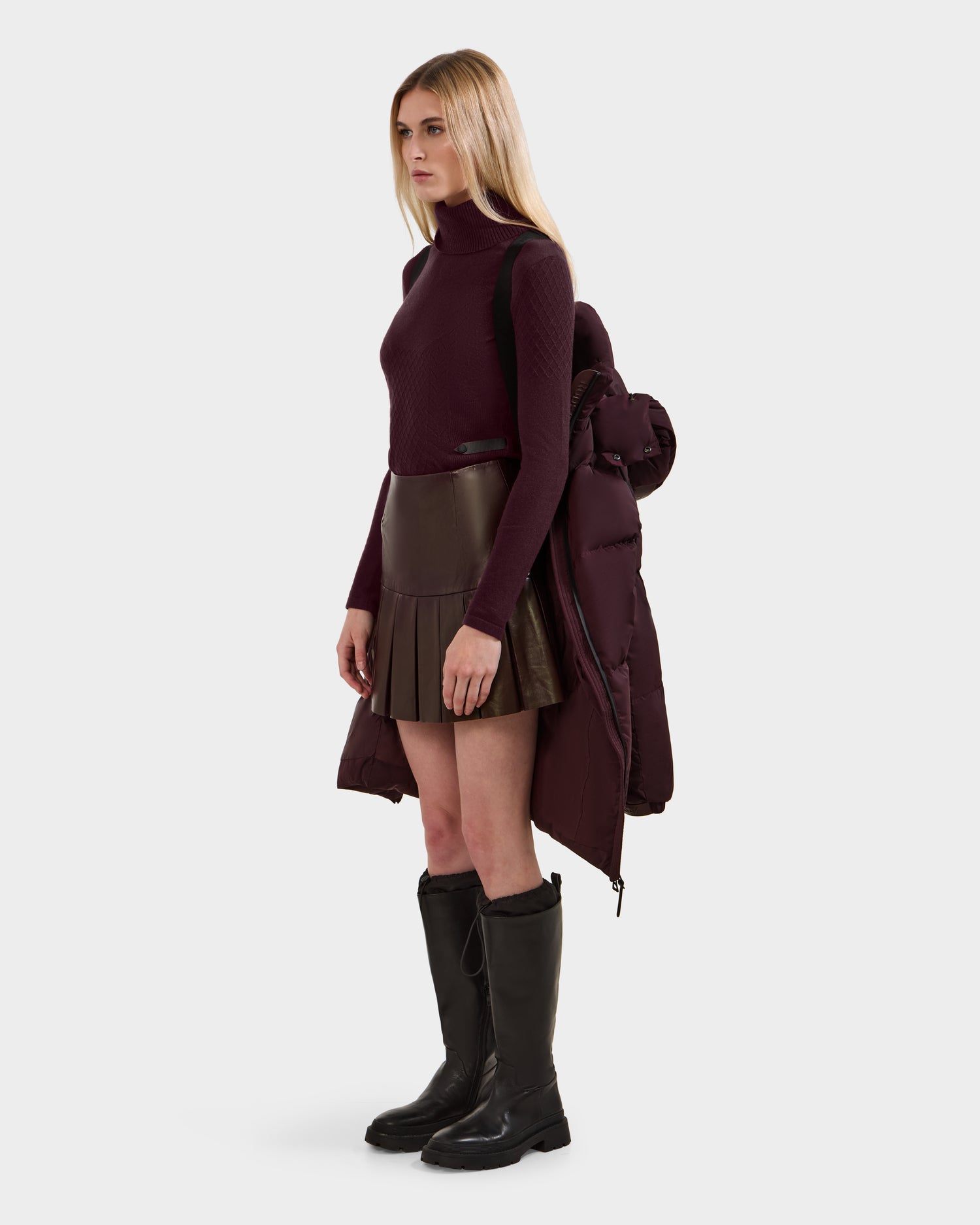 A woman with long blonde hair in a burgundy turtleneck, brown mini skirt, and tall black boots stands against a light background, carrying the Rudsak KARLEE L - 8125682 Bordeaux women's long puffer with vegan leather trim over her shoulders.