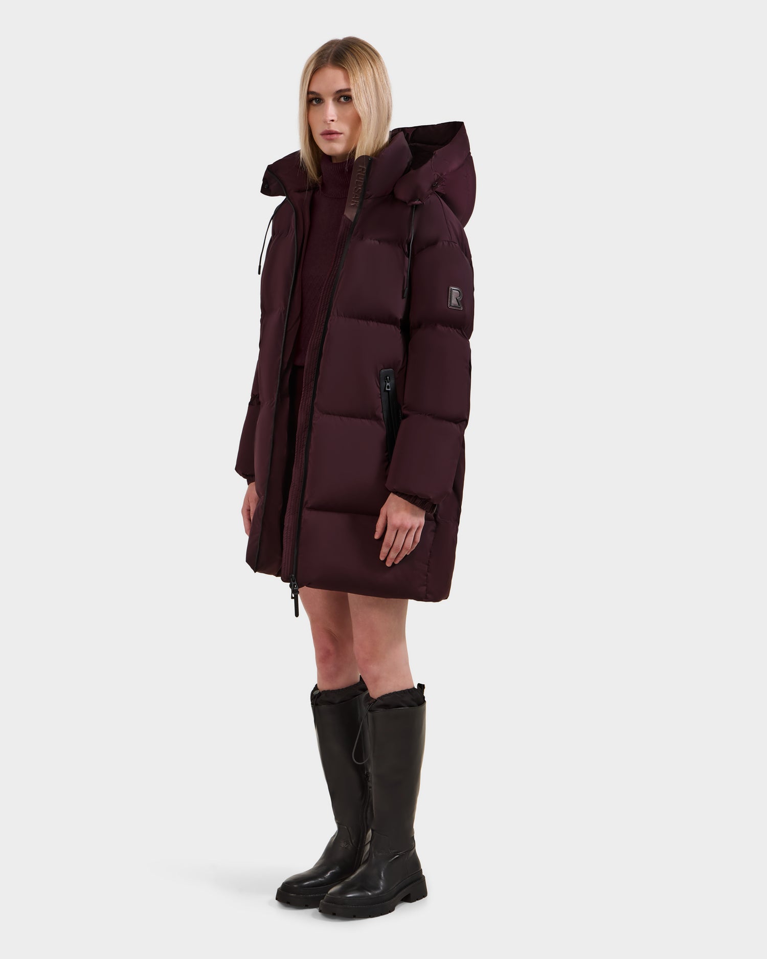 A woman with straight, shoulder-length blonde hair and a neutral expression wears the Rudsak KARLEE L - 8125682 Bordeaux women's long puffer down winter jacket, paired with a matching sweater and knee-high black boots, against a plain background.