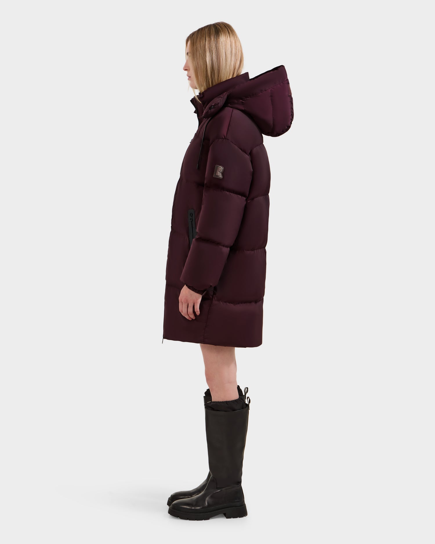 A woman stands in profile wearing the Rudsak KARLEE L - 8125682 Bordeaux women’s puffer coat with vegan leather trim and black knee-high boots against a plain white background.
