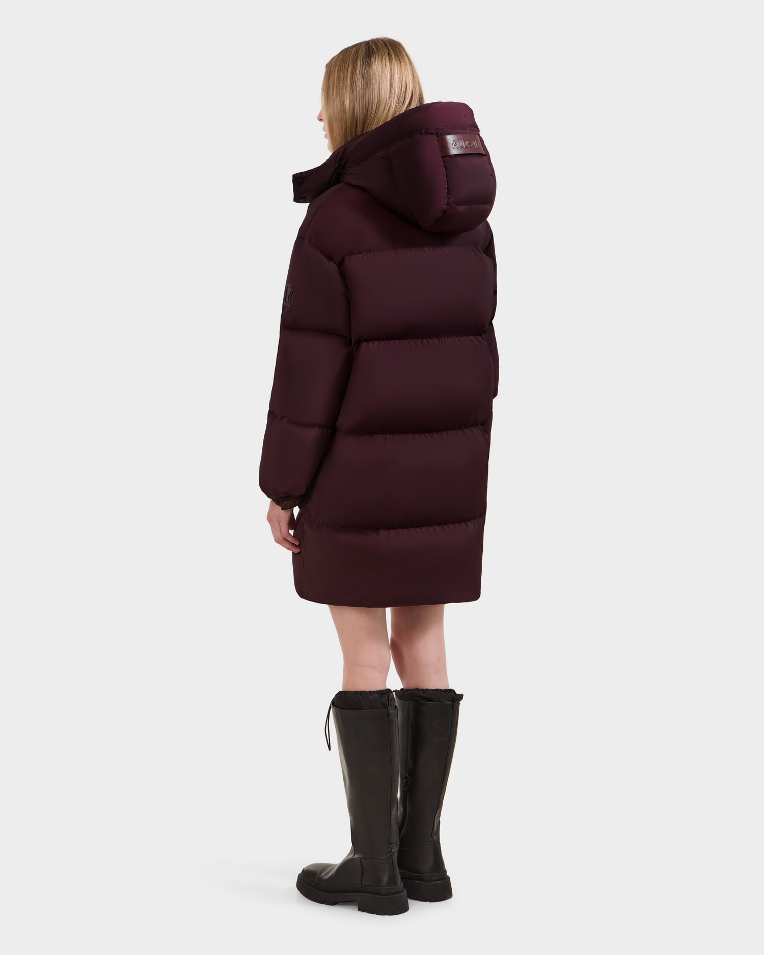 A person with light hair wears the Rudsak KARLEE L - 8125682 Bordeaux women's long puffer, paired with black knee-high boots, standing and facing away from the camera against a plain white background.