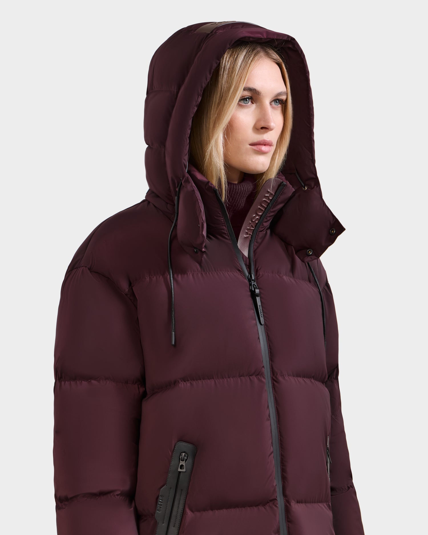 A woman in the Rudsak KARLEE L - 8125682 Bordeaux long puffer jacket with vegan leather trim stands against a plain light background, gazing to the side with a neutral expression.