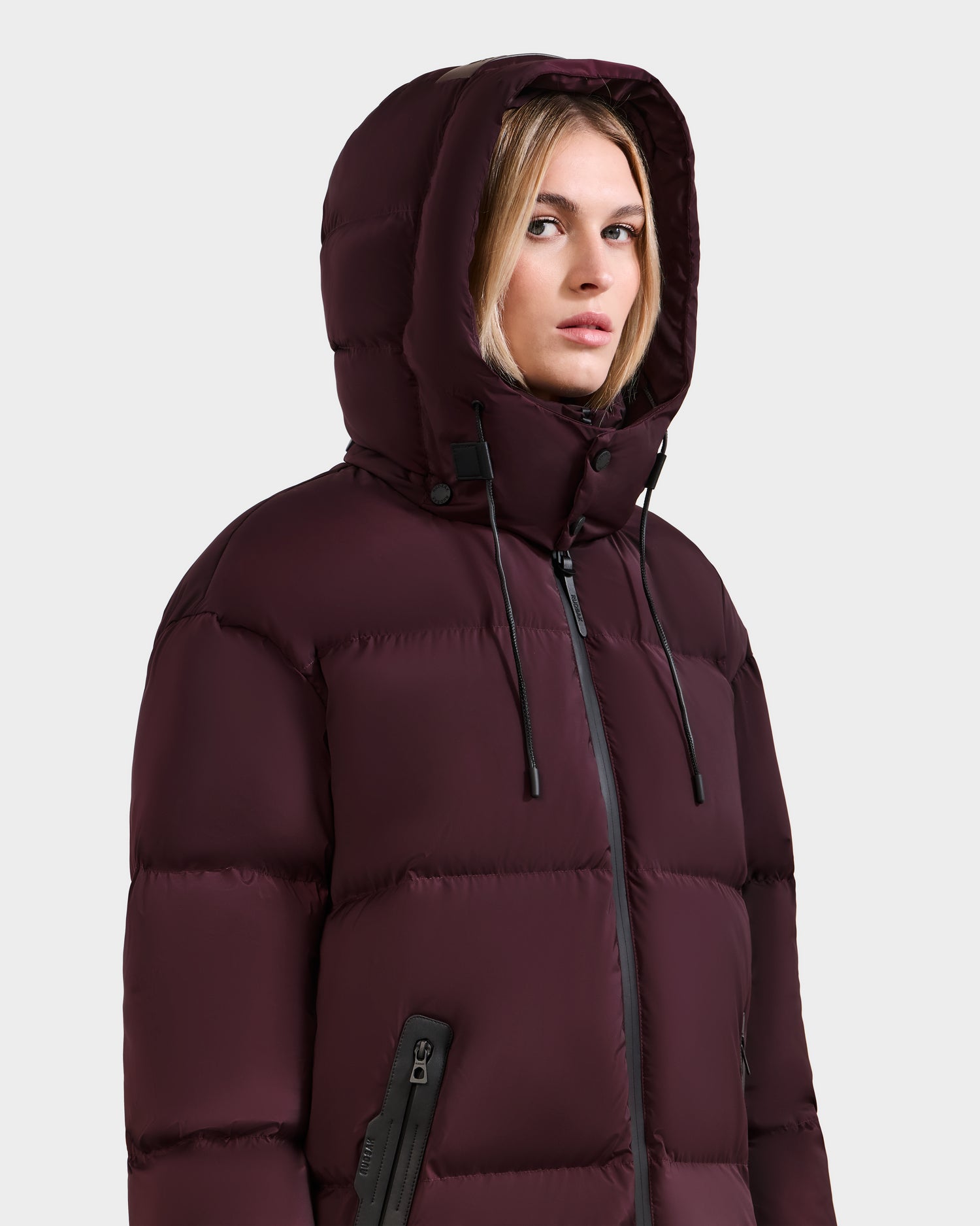 A woman wears the Rudsak KARLEE L - 8125682 Bordeaux, a maroon long puffer jacket with black hood drawstrings, a zippered sleeve pocket, and vegan leather-trimmed front zipper, looking slightly to the side against a plain light background.