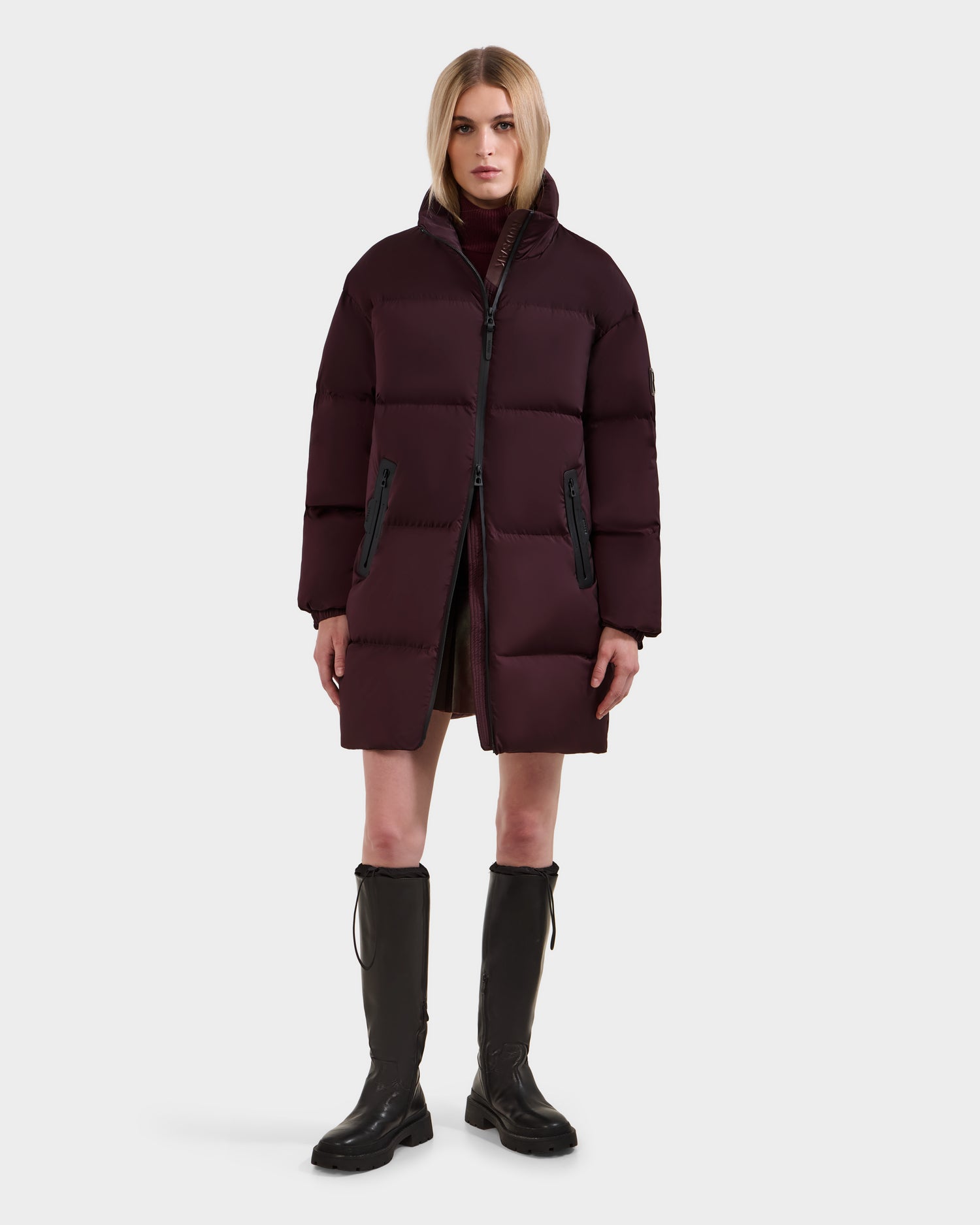 A person stands facing forward against a plain light background, wearing Rudsak’s KARLEE L - 8125682 Bordeaux women’s long puffer coat with vegan leather trim, a high collar, a burgundy turtleneck, and knee-high black boots.