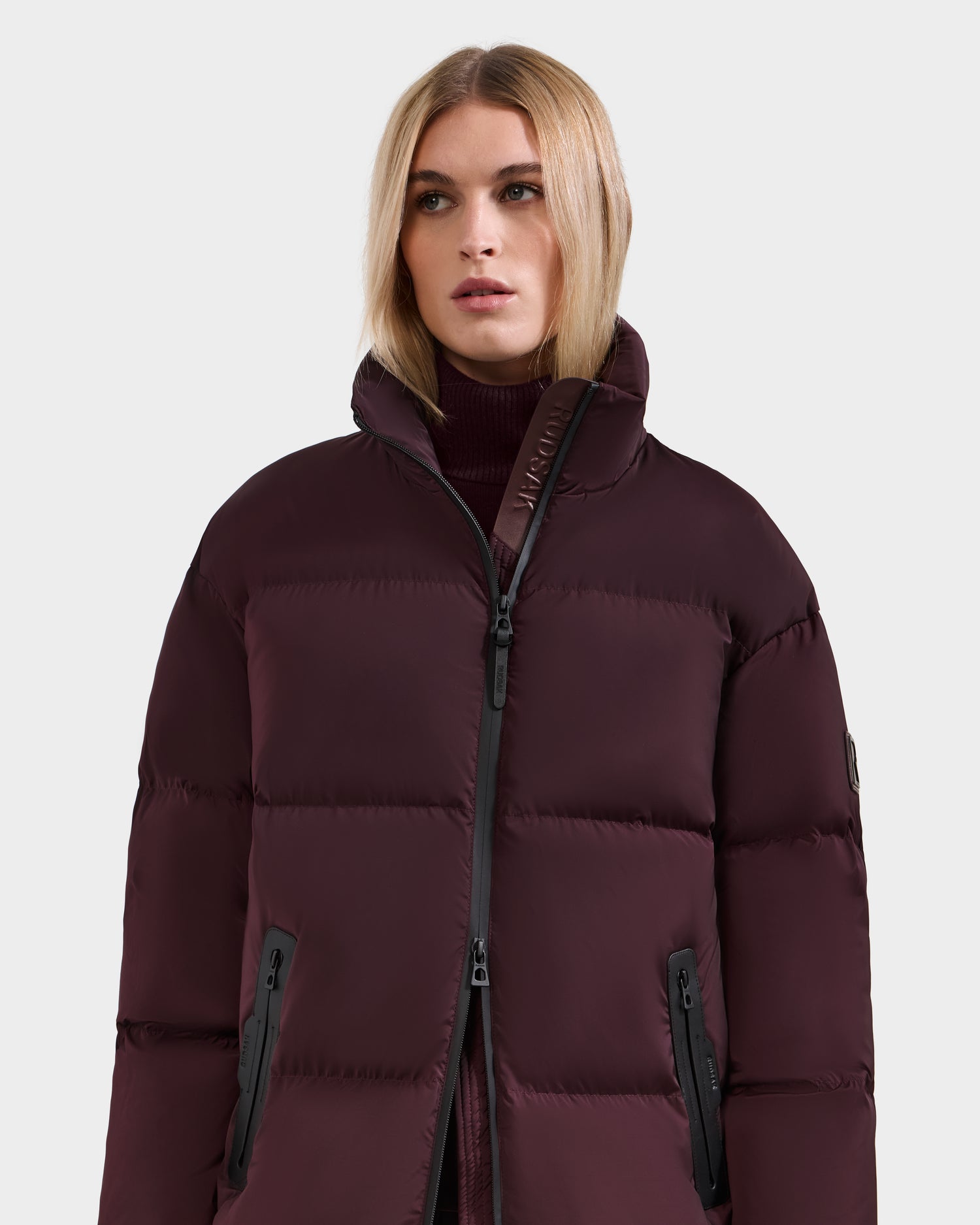 A woman with straight blonde hair models the Rudsak KARLEE L - 8125682 Bordeaux long puffer down winter jacket with a high collar and zippered pockets, set against a plain light gray background.