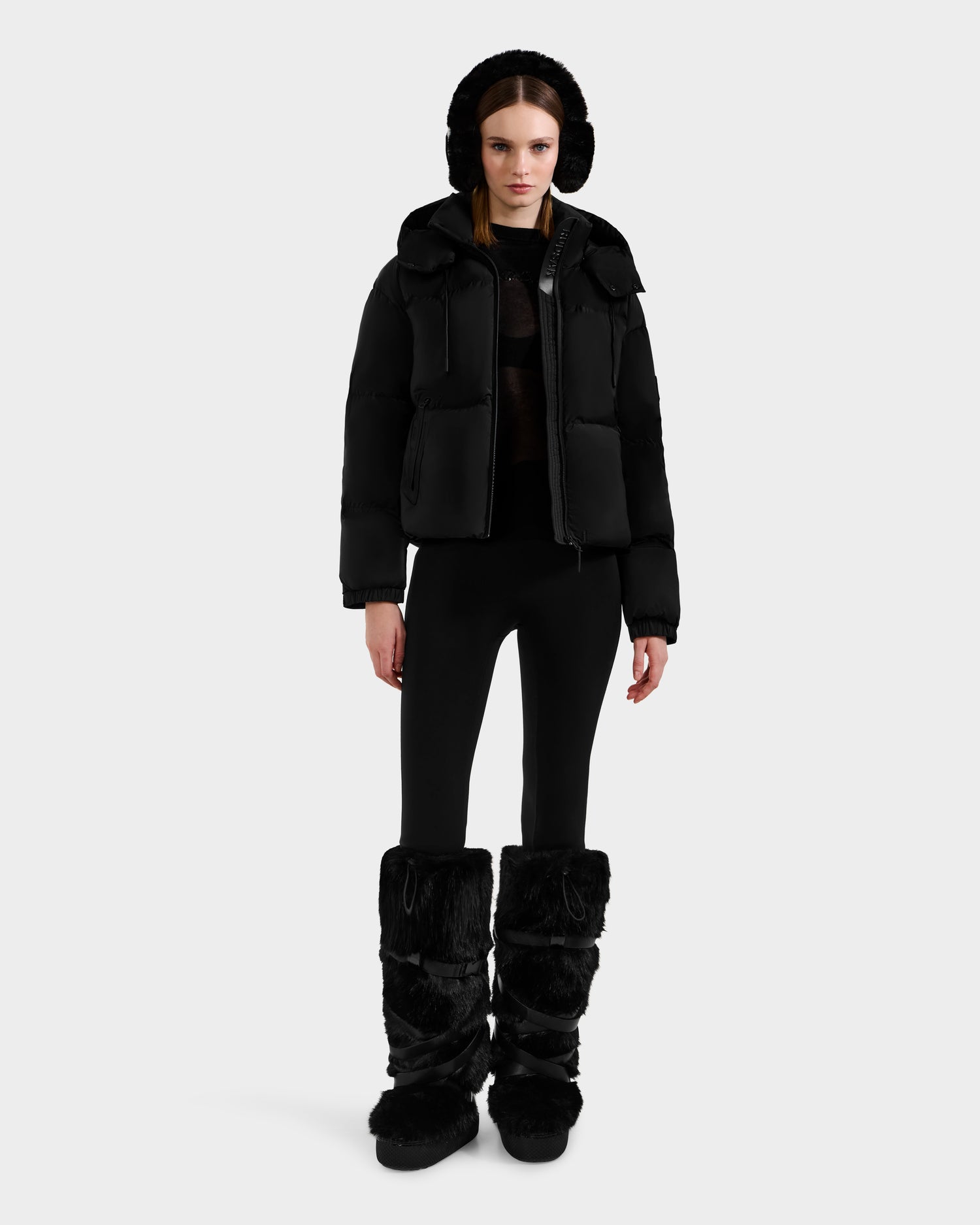 A woman faces forward in a Rudsak KARLEE S - 8125699 Black women’s down puffer jacket with a hood, black leggings, and furry boots, standing against a plain white background.