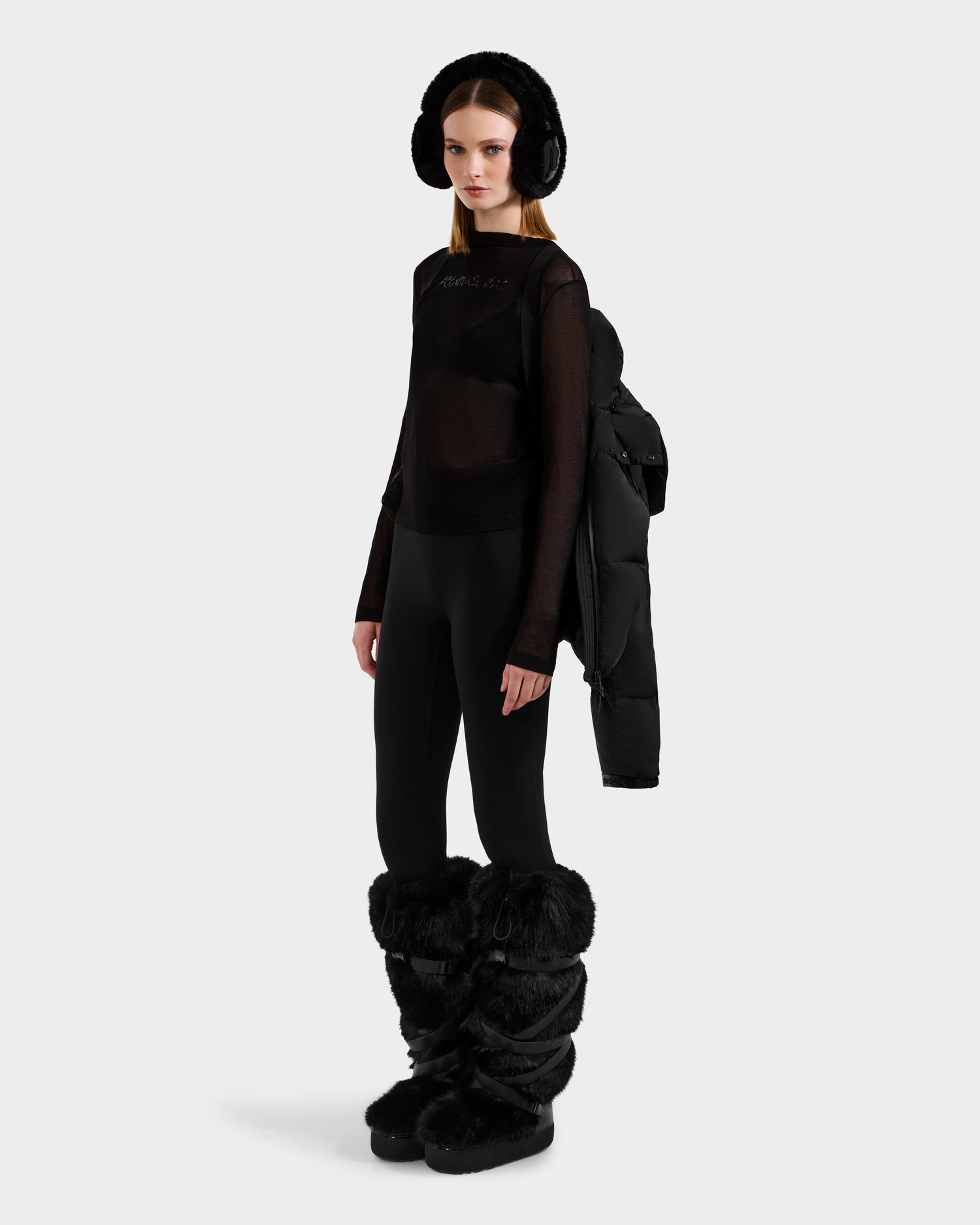 A woman poses against a white background wearing black leggings, a sheer black long-sleeve top, furry black earmuffs and boots, with a Rudsak KARLEE S - 8125699 Black women’s down puffer jacket draped over one shoulder.