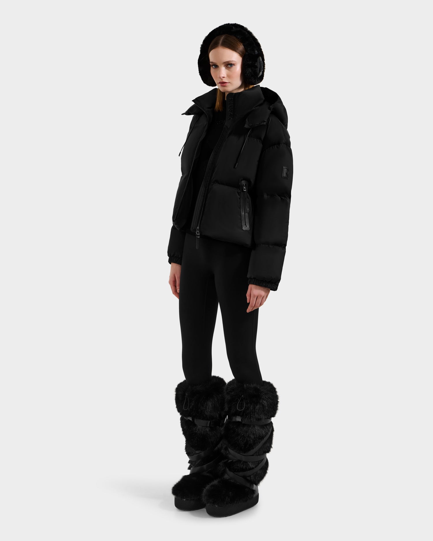 A woman wears the Rudsak KARLEE S - 8125699 Black down puffer jacket with a hood, paired with black leggings and tall black fur boots, standing against a plain white background and looking straight ahead.