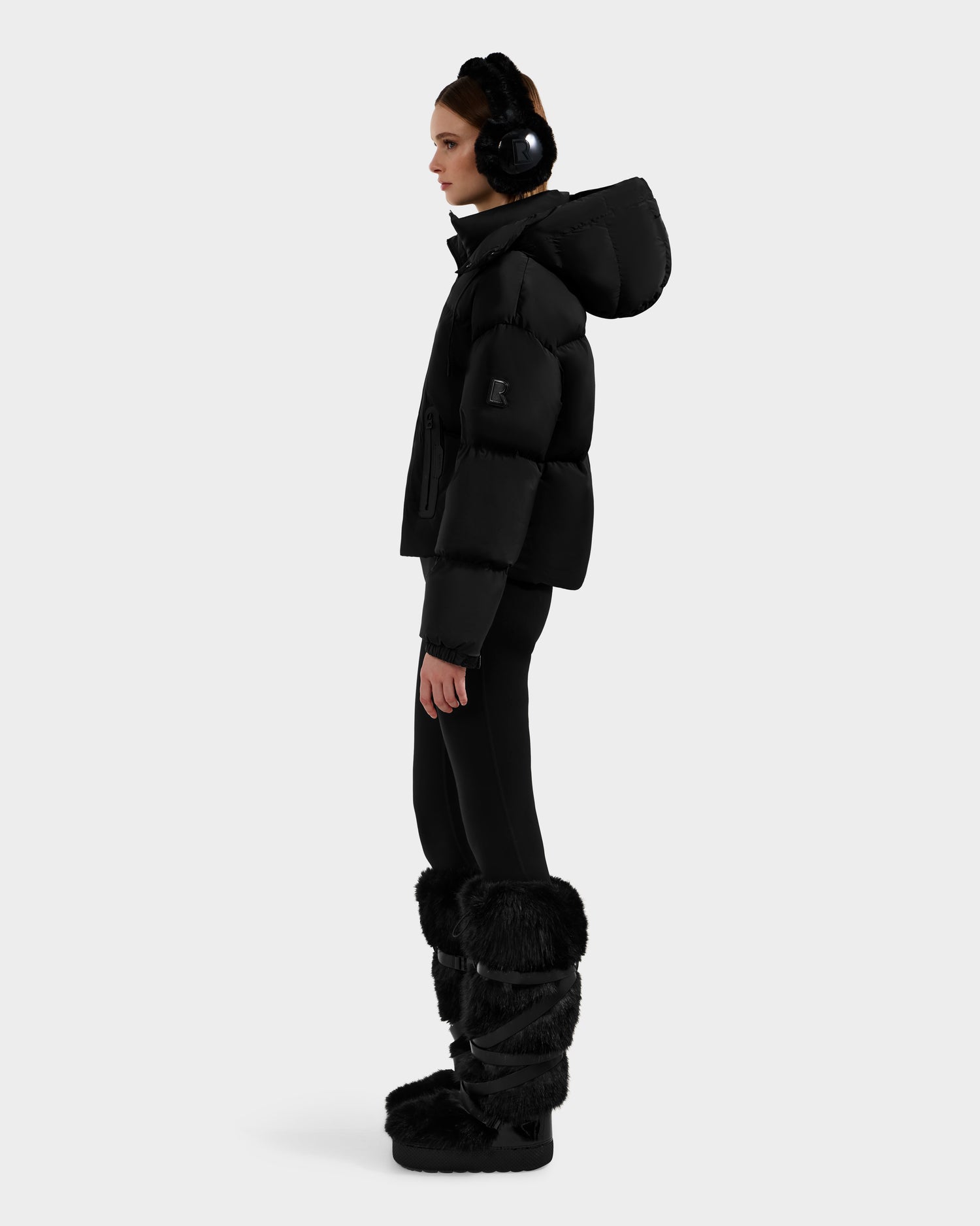 A woman stands in profile wearing the Rudsak KARLEE S - 8125699 Black women's down puffer jacket, styled with black earmuffs, leggings, and tall furry boots, set against a plain light gray background.
