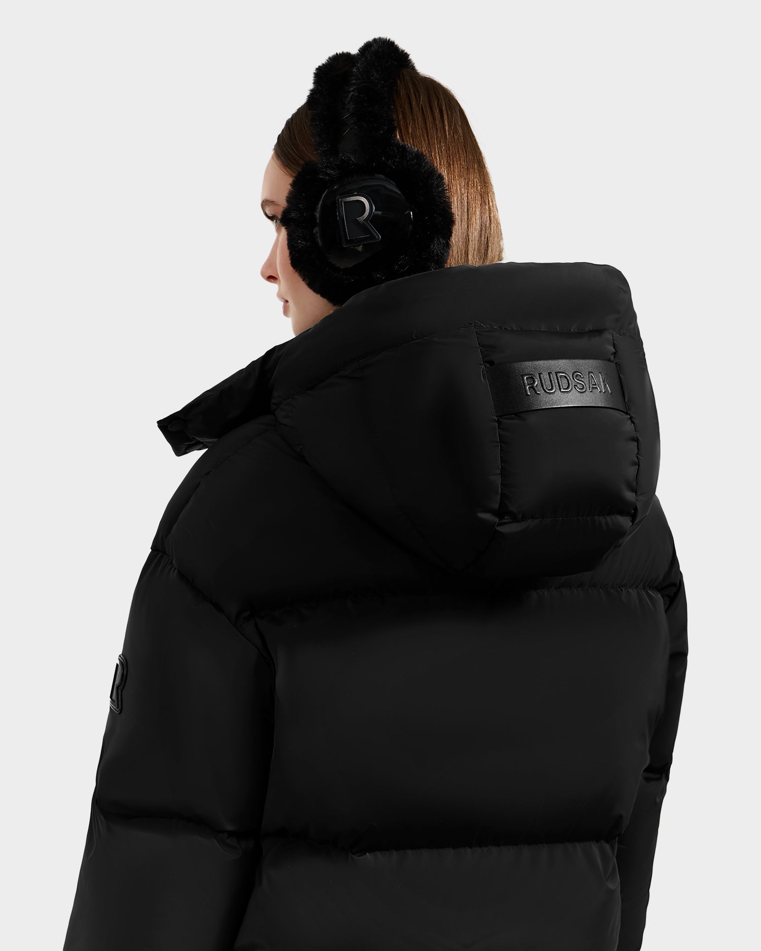 A woman with brown hair faces away from the camera, wearing black earmuffs and the Rudsak KARLEE S - 8125699 women’s down puffer jacket with the logo on the back, set against a plain light background.