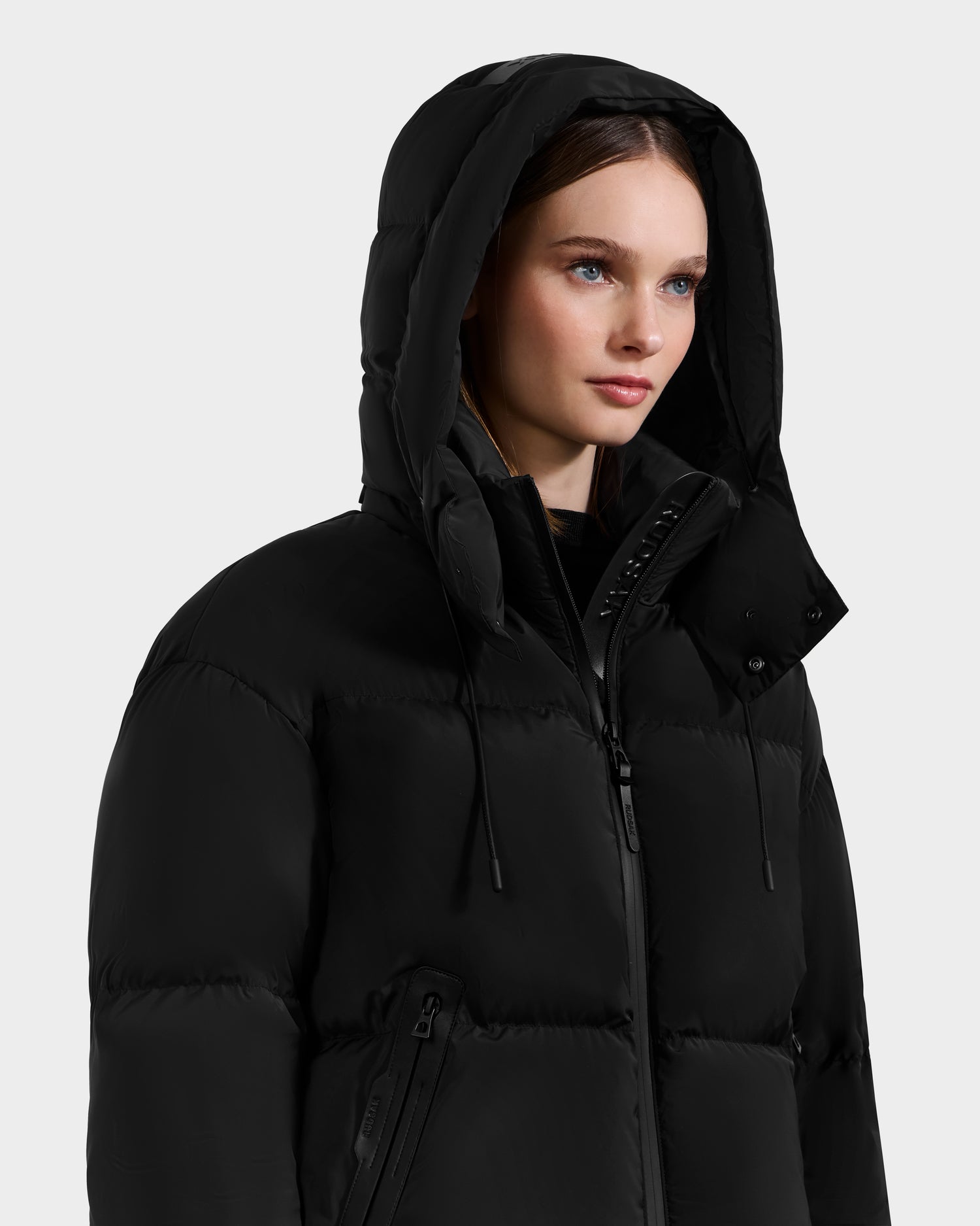 A person wearing the Rudsak KARLEE S - 8125699 Black women’s down puffer jacket with the hood up, looking slightly to the side against a plain light background.
