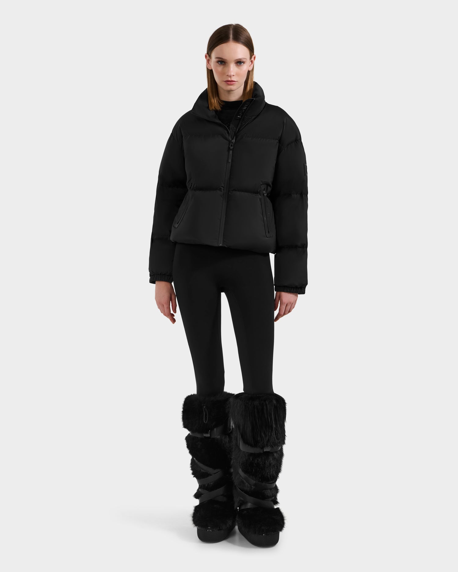 A woman faces forward against a white background, wearing the Rudsak KARLEE S - 8125699 Black women’s down puffer jacket, paired with black leggings and large black fur-trimmed winter boots.