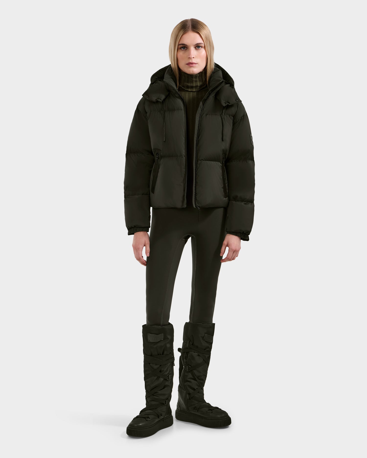 A person with straight blonde hair stands against a plain background, wearing the Rudsak KARLEE S - 8125699 Dark Olive women’s down puffer jacket, black leggings, knee-high quilted boots, and a dark high-neck sweater—a chic cold-weather look.