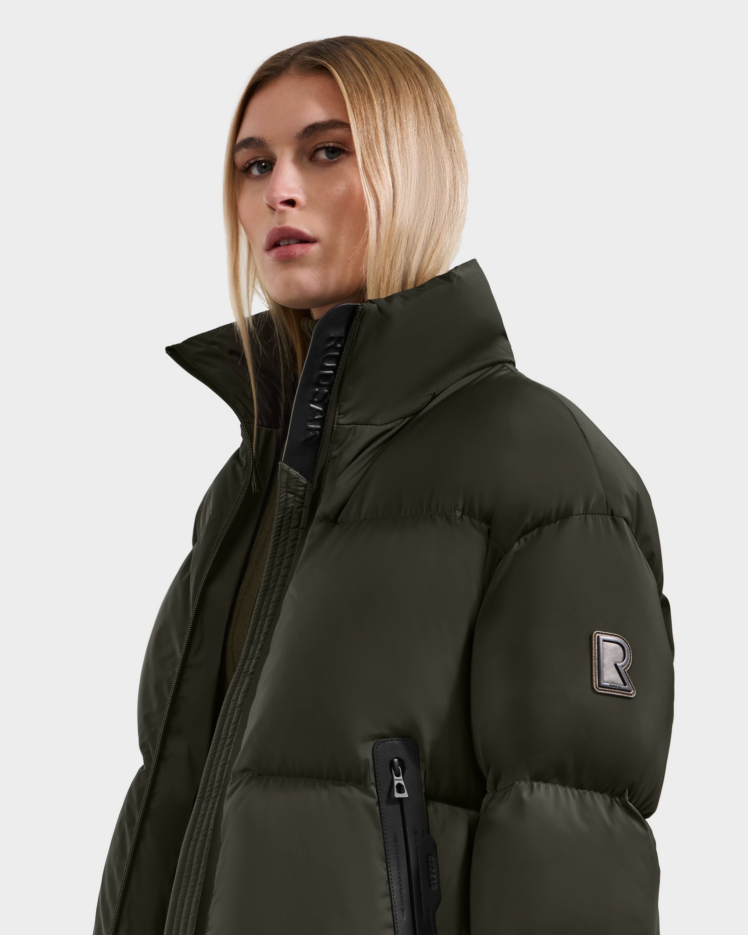 A woman with straight blonde hair wears the Rudsak KARLEE S - 8125699 Dark Olive down puffer jacket with a high collar, visible zipper pocket, and R patch on the sleeve. The background is plain light gray.