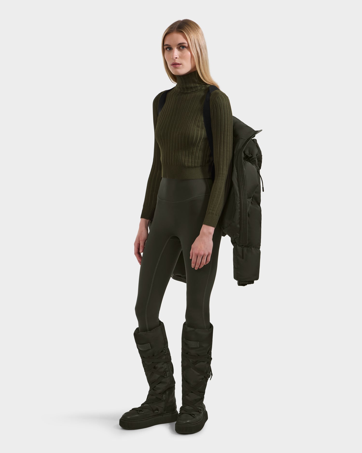 A woman wears the Rudsak KARLEE S - 8125699 Dark Olive down puffer jacket slung over her shoulder, styled with an olive ribbed turtleneck, high-waisted leggings, and quilted knee-high boots against a white background.