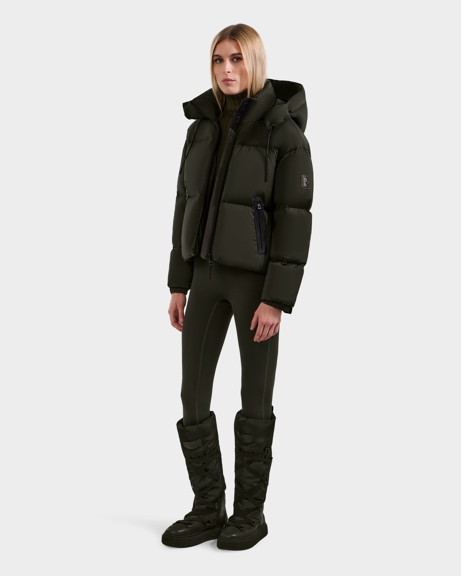 A woman with straight light blonde hair and a neutral expression wears the Rudsak KARLEE S - 8125699 Dark Olive women’s down puffer jacket against a plain background, styled with matching fitted pants and knee-high quilted boots—perfect for cold weather.