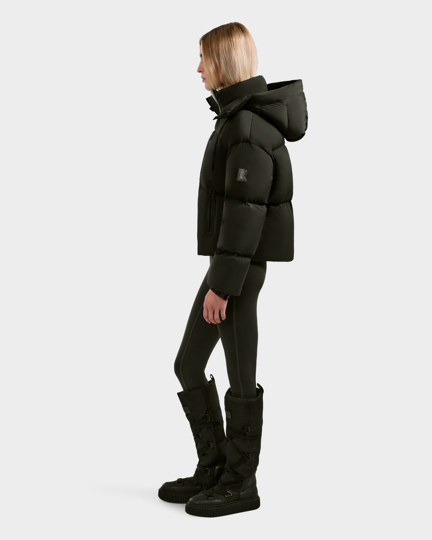 A person stands in profile wearing the Rudsak KARLEE S - 8125699 Dark Olive women’s down puffer jacket with a hood, paired with black leggings and knee-high boots—an ideal cold-weather essential—against a plain light background.