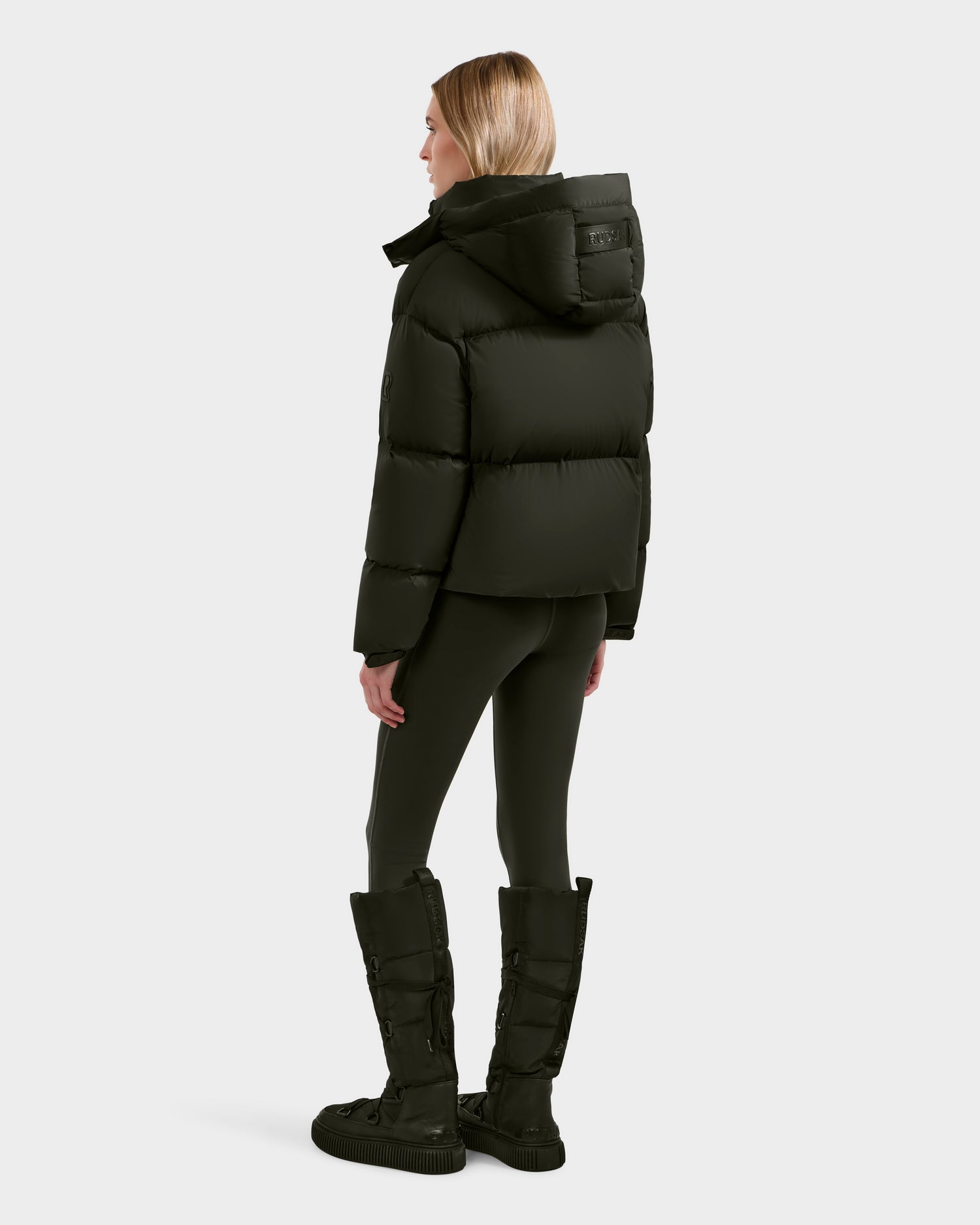 A woman faces away, wearing Rudsak's KARLEE S - 8125699 Dark Olive down puffer jacket with a hood, black leggings, and tall quilted boots—her straight blonde hair stands out against the light gray background in this cold-weather look.