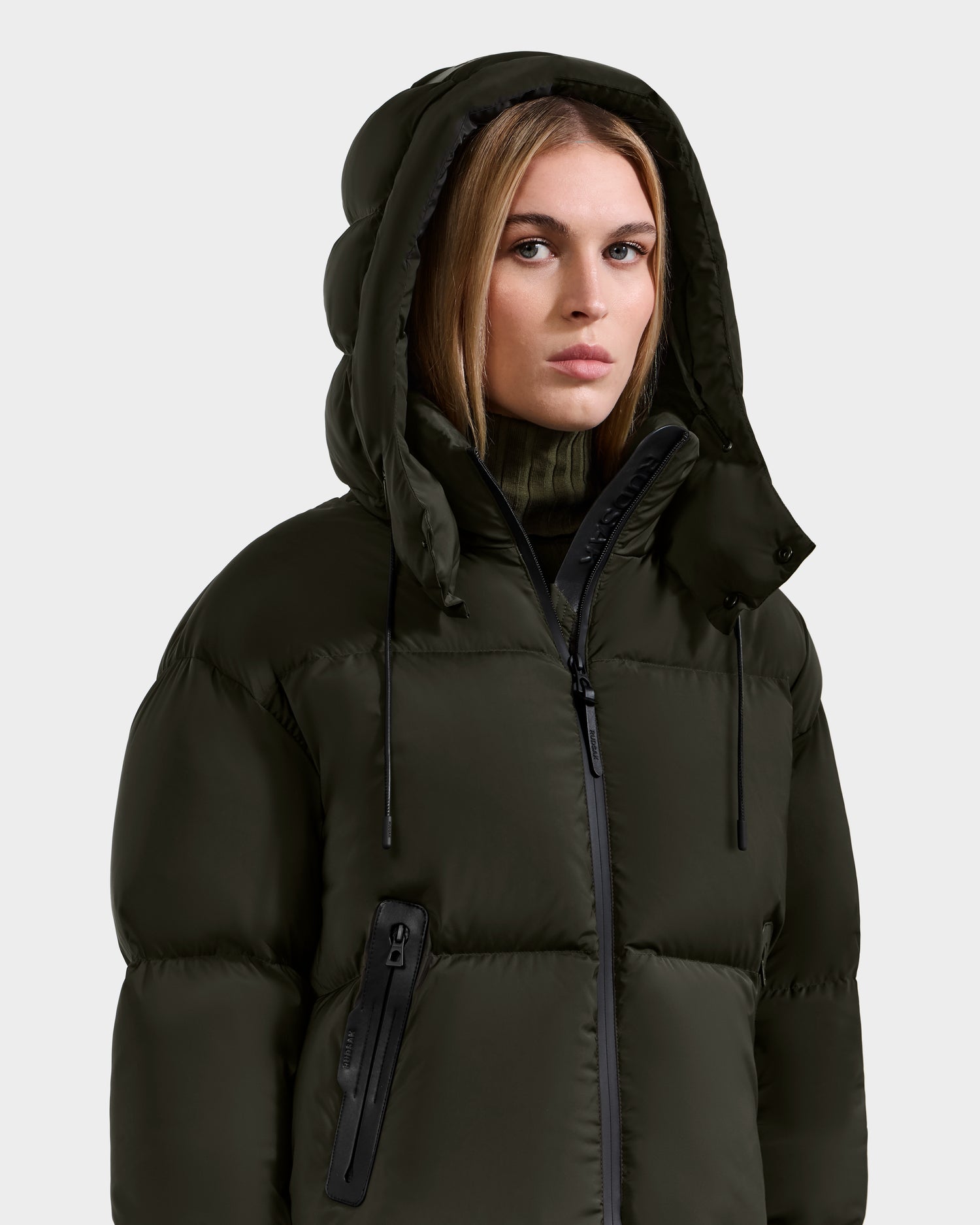 Wearing the Rudsak KARLEE S - 8125699 Dark Olive women’s down puffer jacket and a high-neck sweater, she faces forward with a neutral expression against a plain light background—embodying cold-weather essentials.