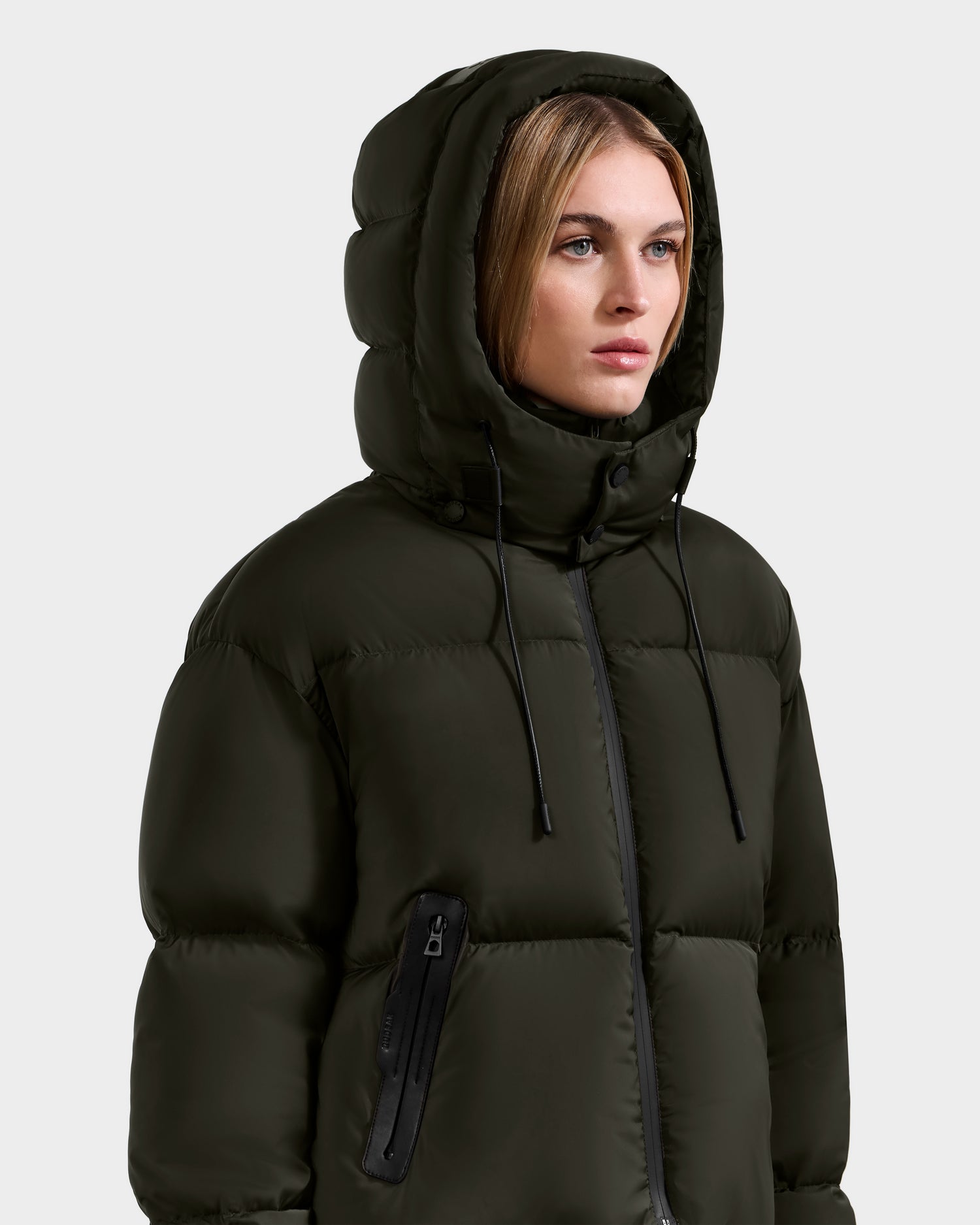 A woman wearing the Rudsak KARLEE S - 8125699 Dark Olive down puffer jacket with hood up, looking to the side. This jacket features a chest zipper pocket and black drawstrings on the hood, perfect for cold weather.