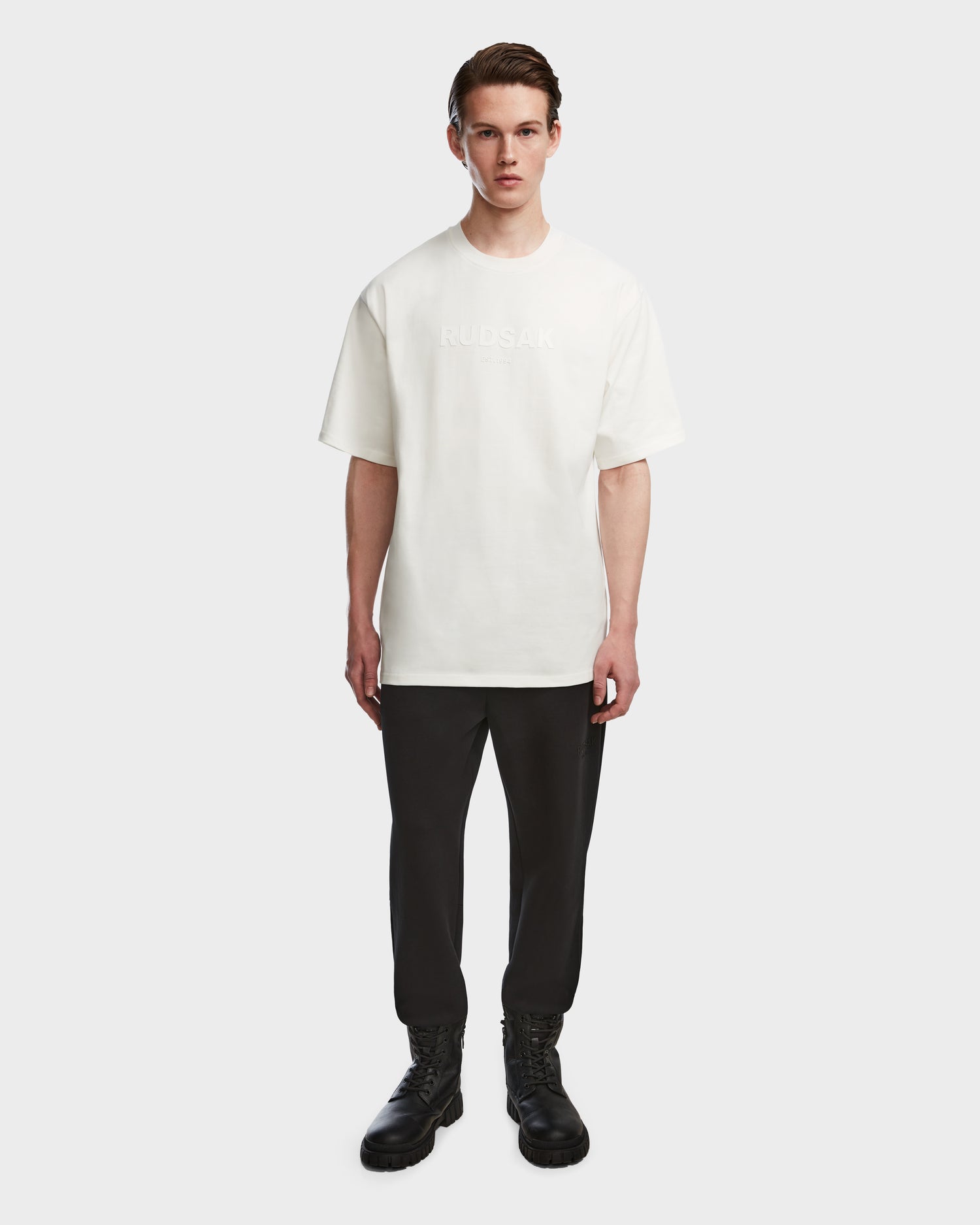 A young man stands facing forward, wearing the Rudsak KARTER - 9524008 Iceland M relaxed fit oversized unisex T-shirt in white, paired with black pants and black lace-up boots against a plain white background.