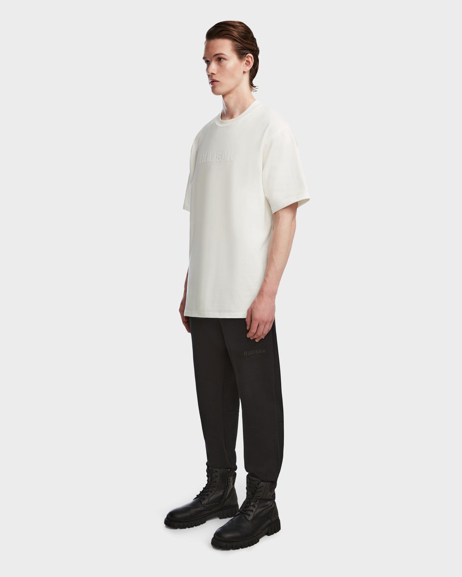 A man faces slightly right with a neutral expression, wearing Rudsaks KARTER - 9524008 Iceland M oversized unisex T-shirt, relaxed fit black pants, and black lace-up boots against a plain background.