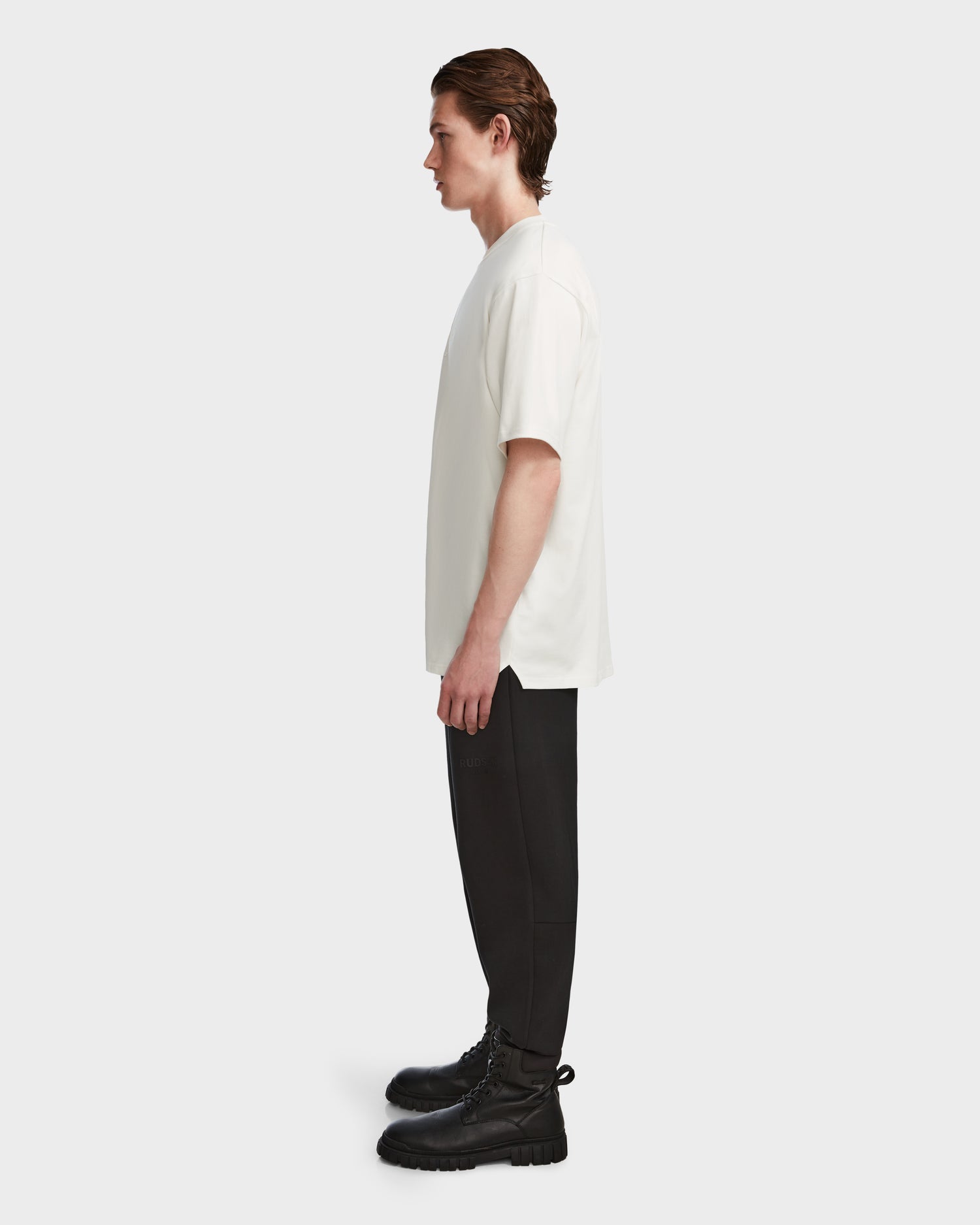A man stands in profile against a plain background, wearing the KARTER - 9524008 Iceland M T-shirt from Rudsak, paired with black pants and black lace-up boots.