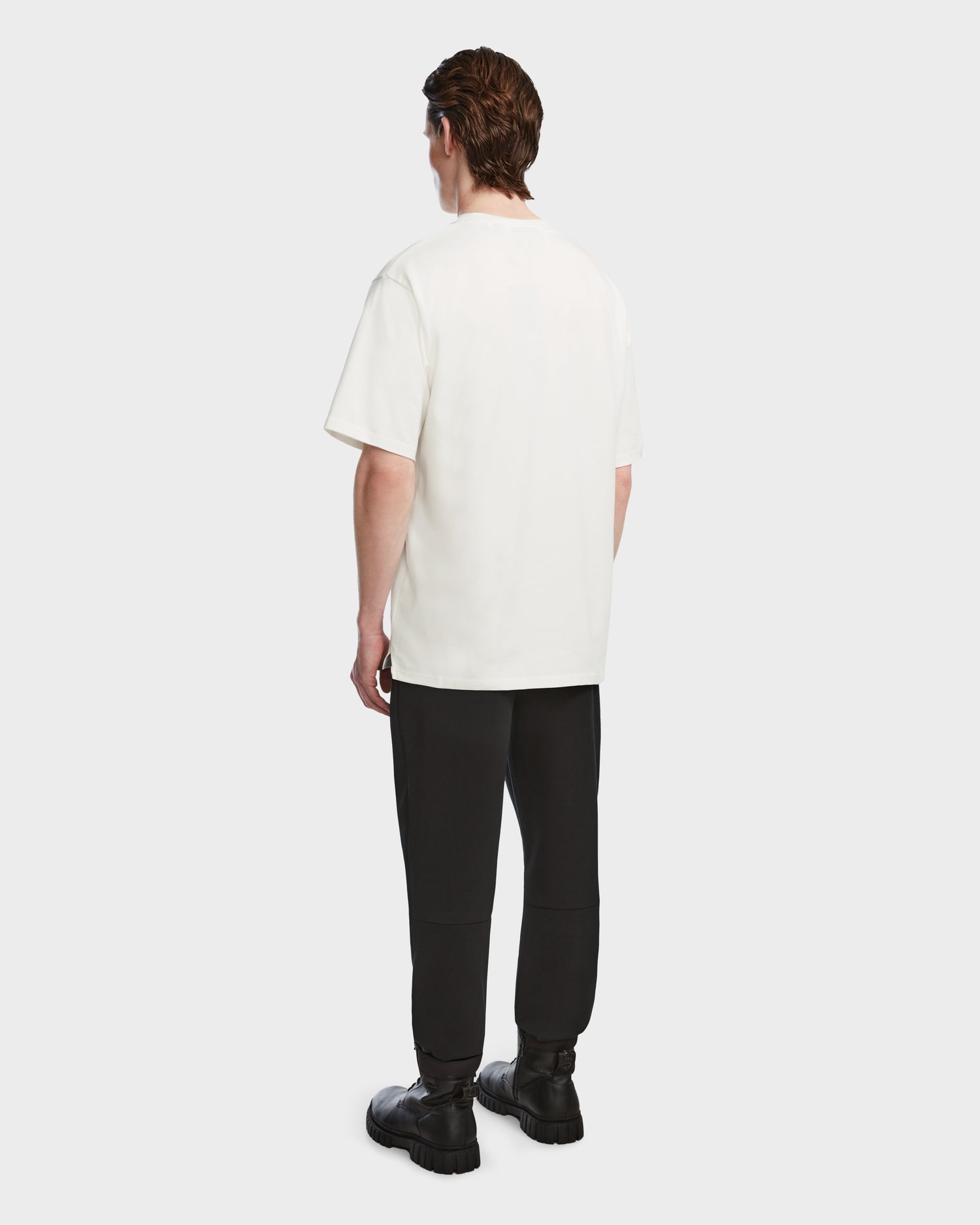 A person faces away, wearing the Rudsak KARTER - 9524008 Iceland M premium cotton T-shirt, black pants, and chunky black boots against a plain light background.