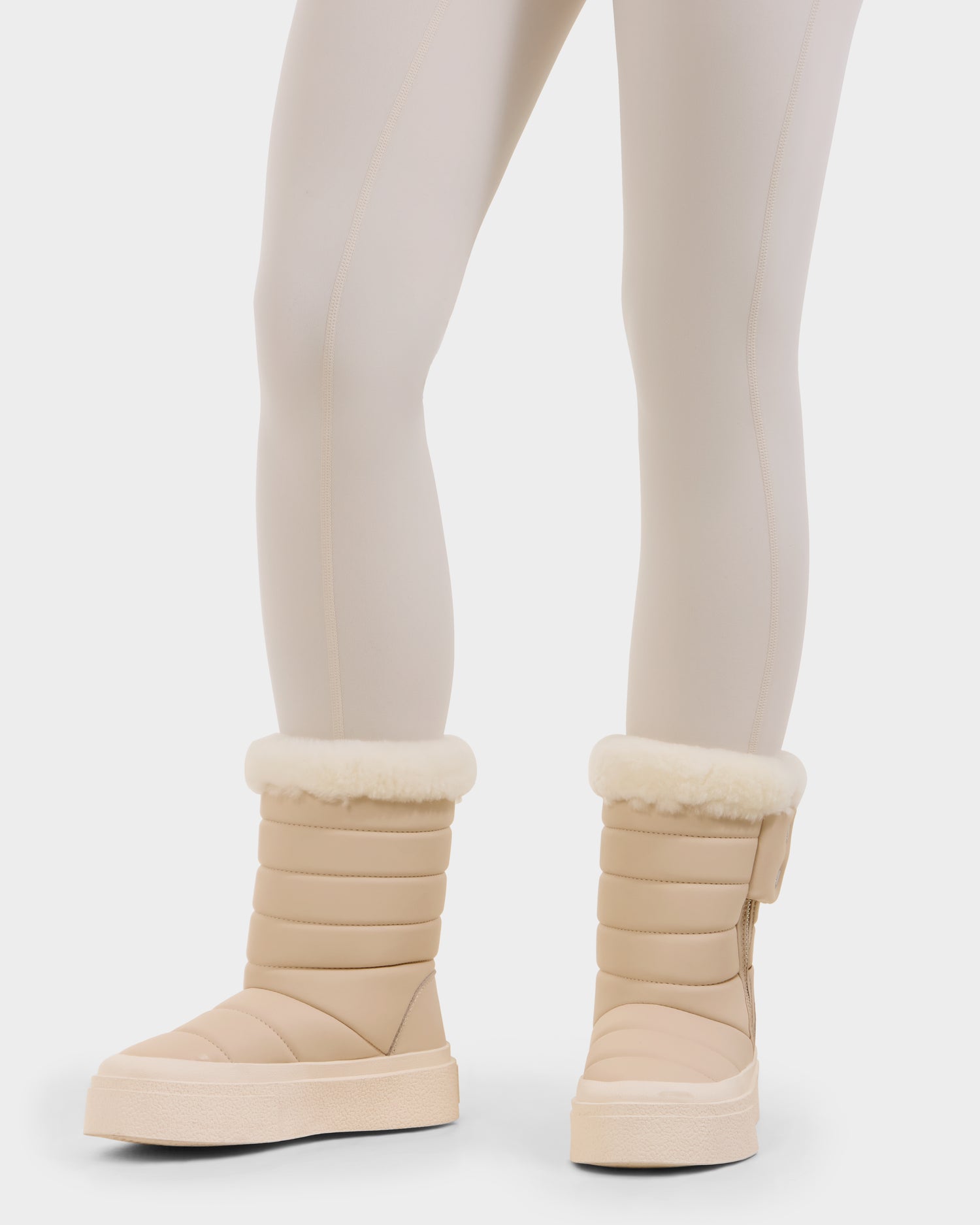 A person wearing light beige leggings and Rudsak KATHIE - L225516 Vanilla vegan leather winter boots with cream-colored faux fur lining stands against a plain white background.