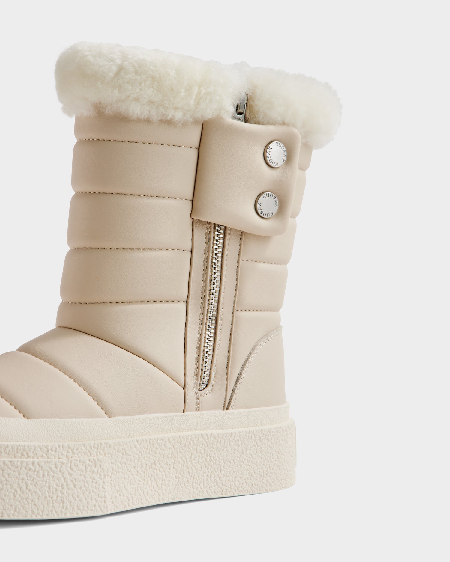 A close-up of the KATHIE - L225516 Vanilla women’s mid boot by Rudsak, featuring beige quilted stitching, a white faux fur cuff, metallic zipper, two silver buttons, and a thick textured sole—perfect for elegant winter style.