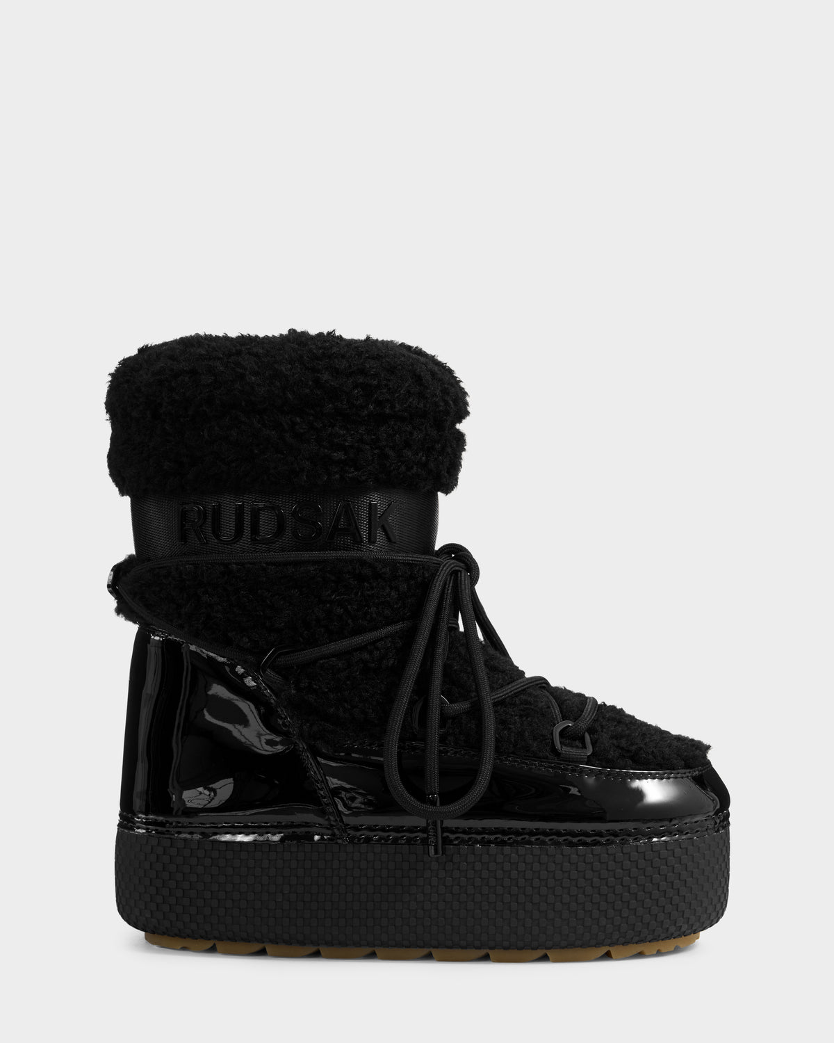 A single KATHY Y - 1224500 Black winter boot by Rudsak features a shiny patent finish, textured sole, thick laces, faux fur upper cuff, and an embossed Rudsak logo on the shaft. The background is plain light gray.