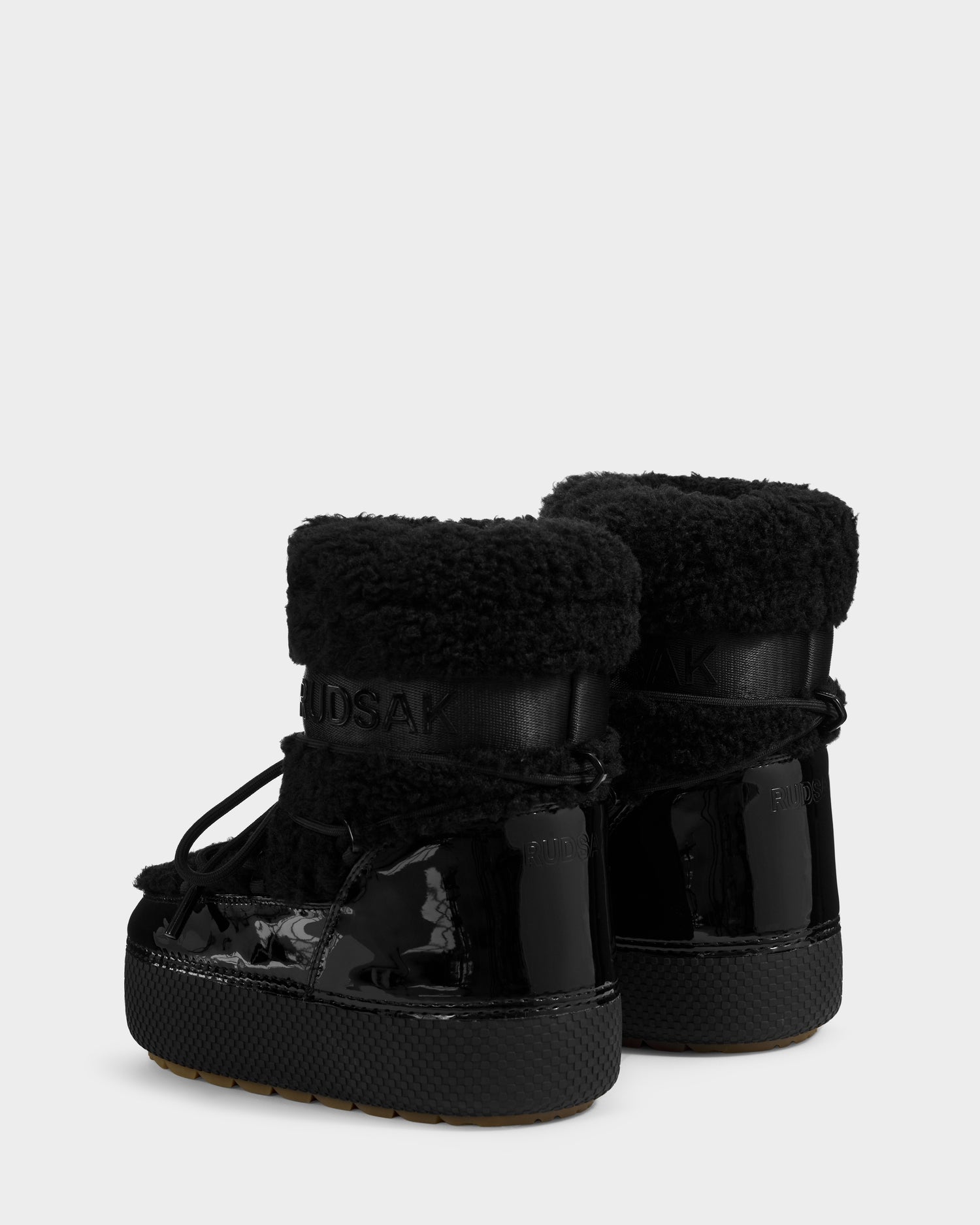 The Rudsak KATHY Y - 1224500 Black boots feature water-repellent shiny patent and faux shearling, thick soles, laces, and raised back cuffs, shown against a plain light gray background.