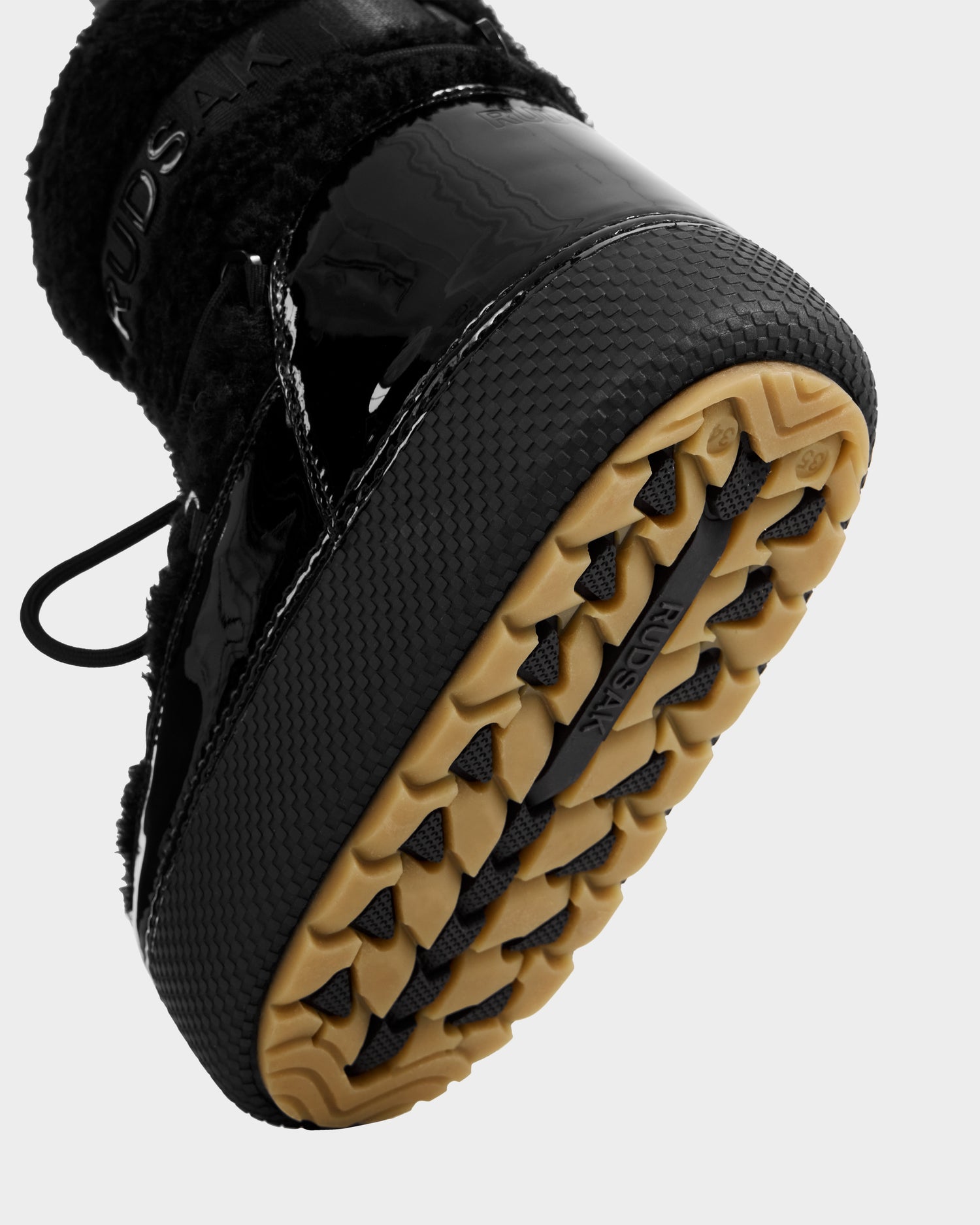 Close-up of the sole of the Rudsak KATHY Y - 1224500 Black water-repellent winter boot, showing a tan, textured rubber outsole, black patent leather, and fuzzy materials on the upper.