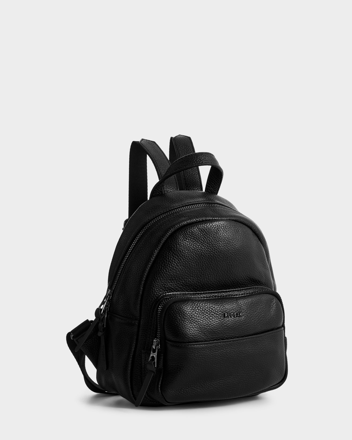 The Rudsak KATTY - L325014 Black mini backpack, made from black pebbled leather, has two zippered compartments, a small front pocket, and adjustable straps. Shown against a plain light gray background.