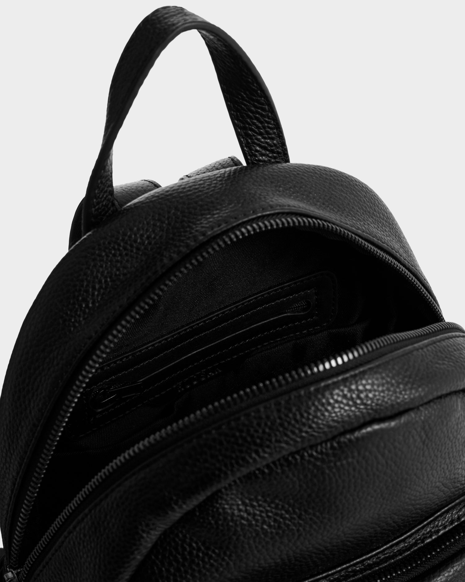 Close-up of the Rudsak KATTY - L325014 Black pebbled leather backpack with a partially open zipper, showing its interior compartments against a plain, light background.