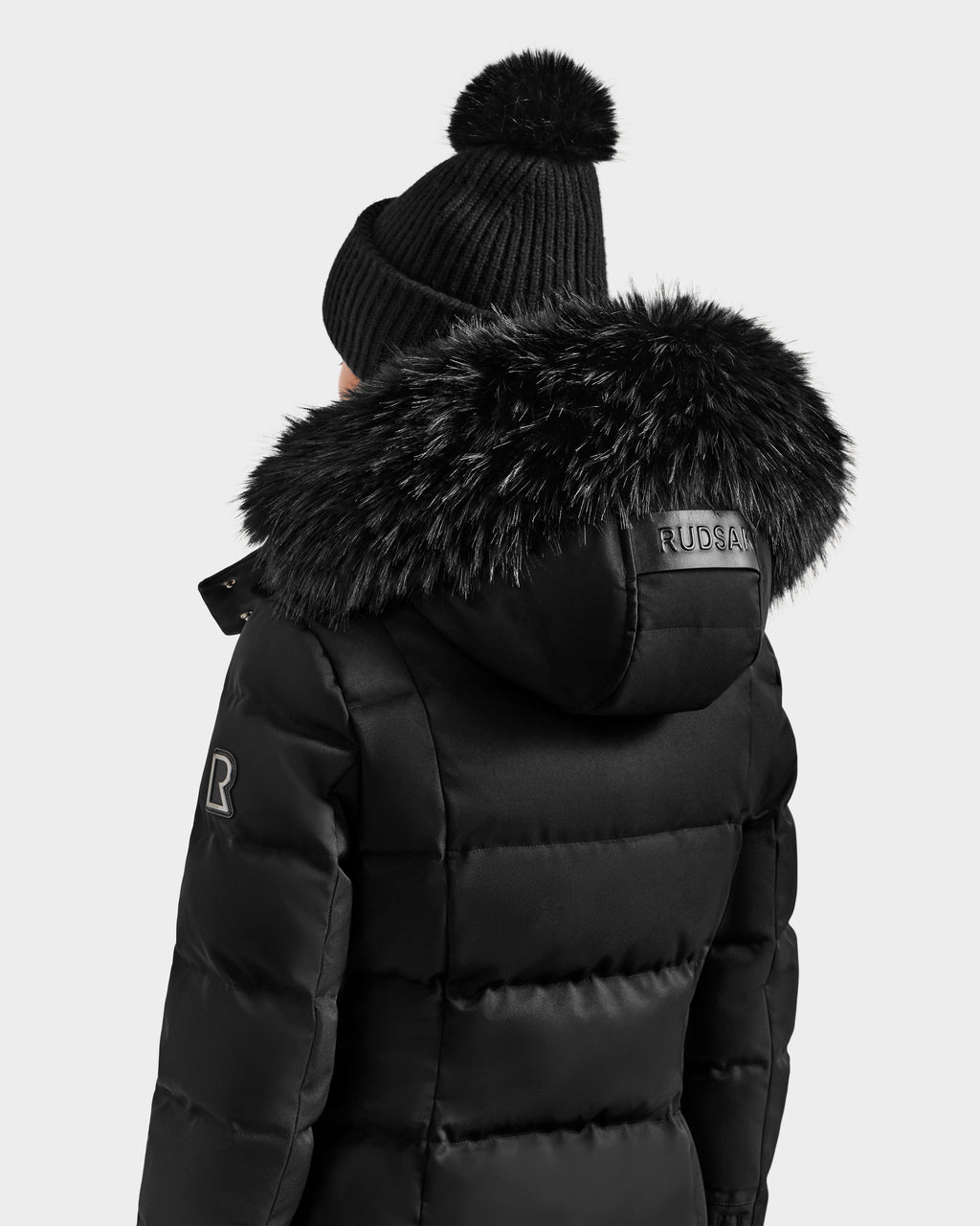 A woman wearing the Rudsak KEHLANI H - 8125643 Black women’s down parka with a faux fur-trimmed hood and a black pom-pom knit beanie, seen from behind against a light background—a true winter essential.