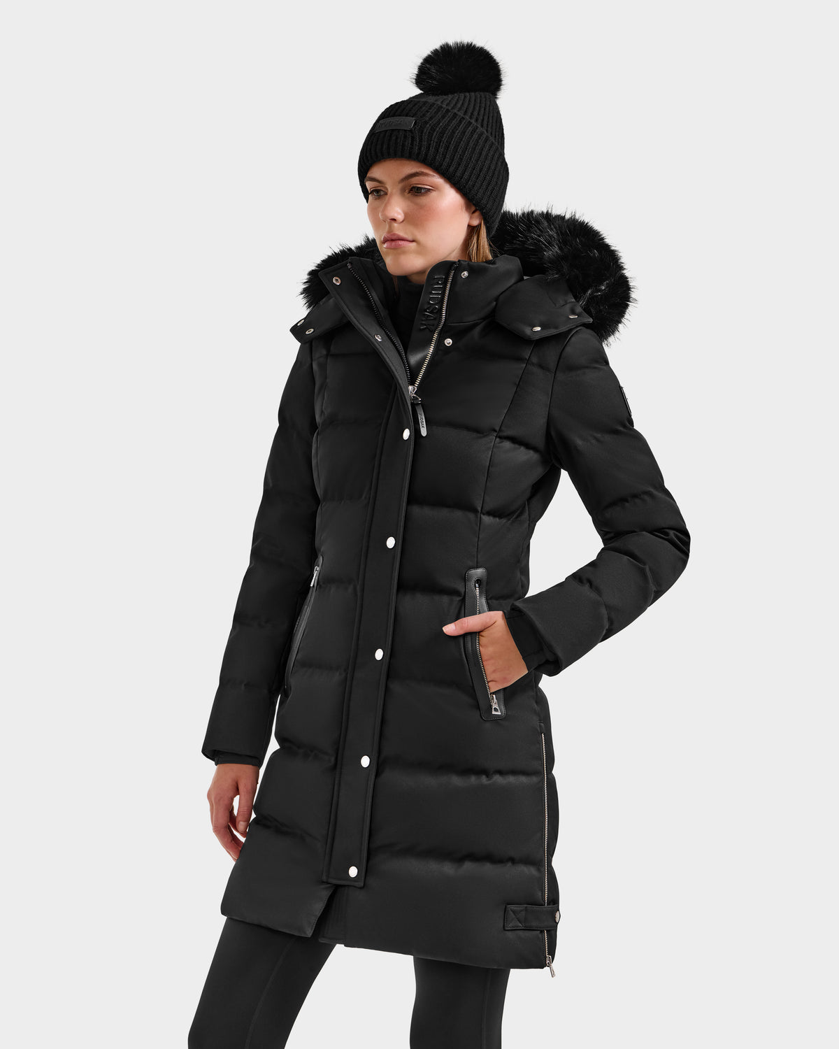 A woman wears the Rudsak KEHLANI H - 8125643 Black down parka with a faux fur-trimmed hood, paired with black leggings and a pom-pom beanie, standing against a plain light background.