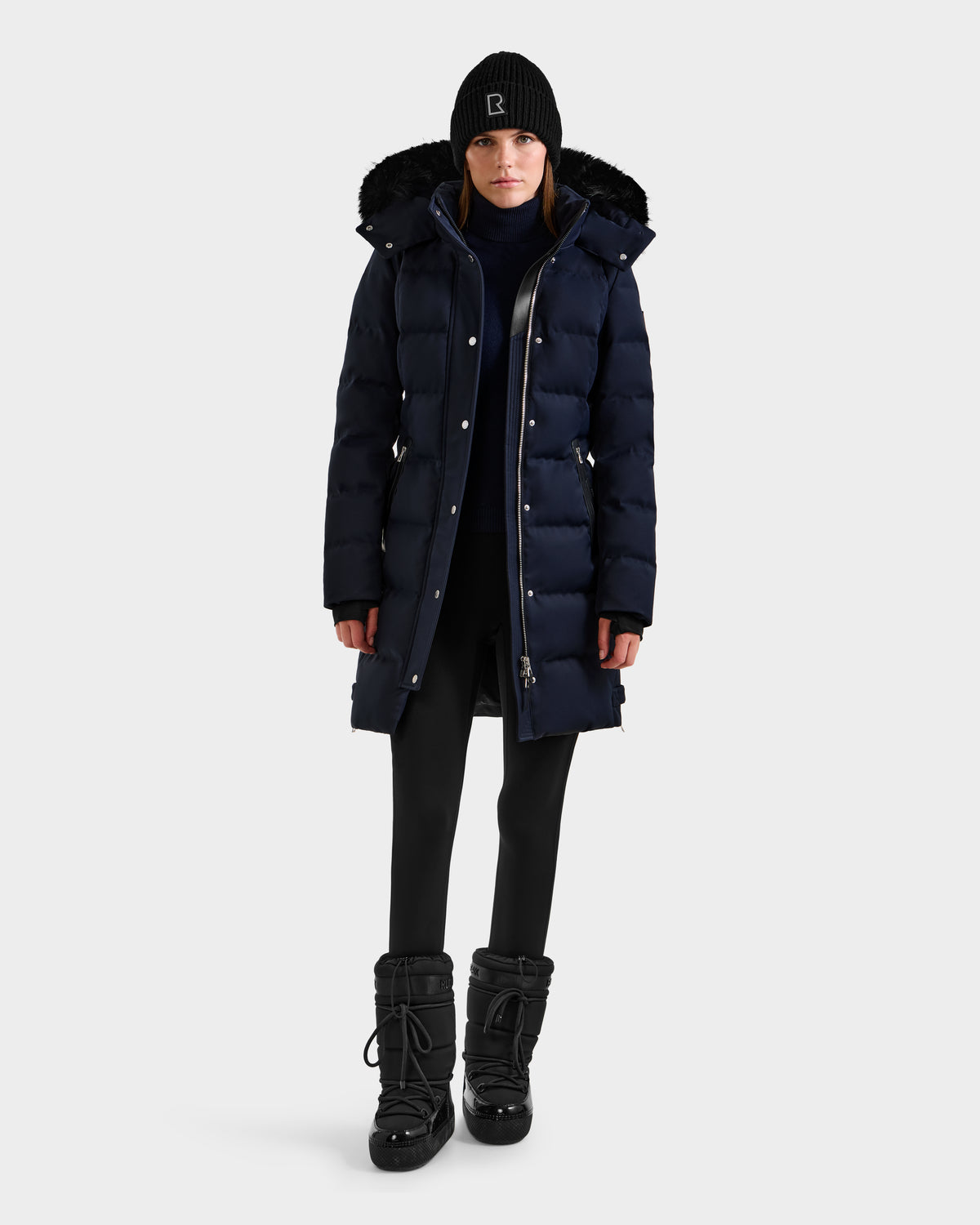 A person wearing the Rudsak KEHLANI H - 8125643 Navy women’s down parka with a fur-lined hood, black leggings, black winter boots, and a black beanie stands against a plain light background. The parka is filled with RDS-certified down.