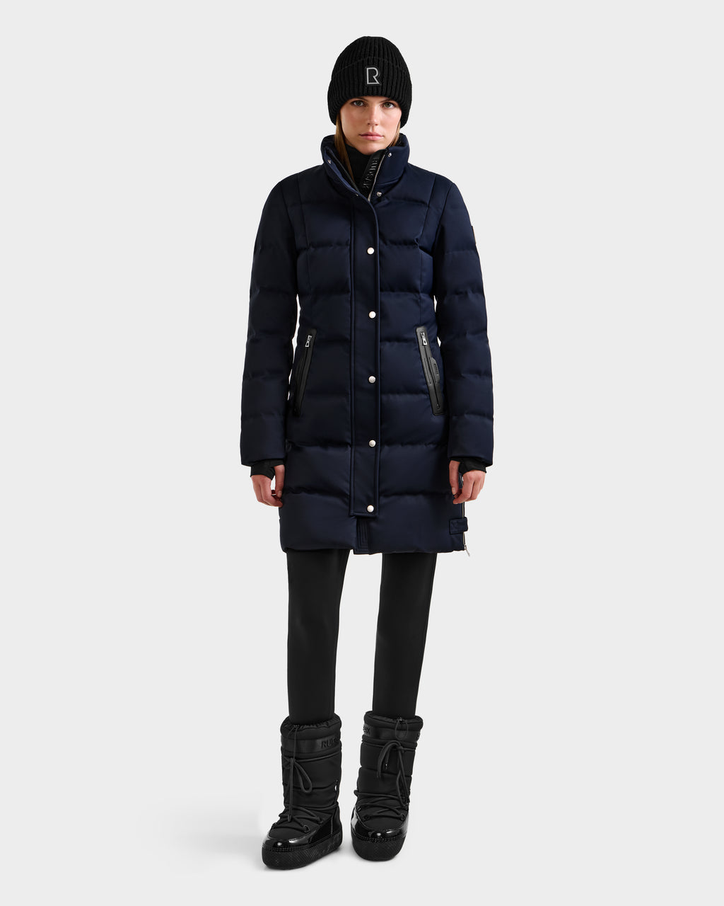 A person stands facing forward against a plain light background, wearing the Rudsak KEHLANI H - 8125643 Navy women’s down parka, paired with black leggings, black winter boots, and a black beanie.