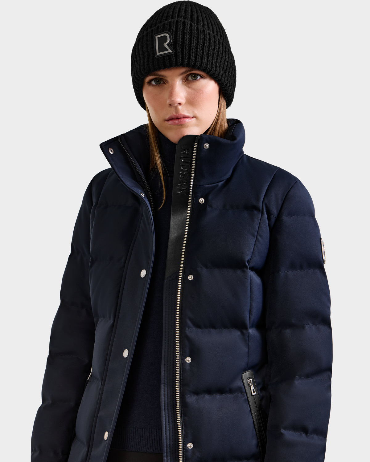 A woman wearing a black knit beanie with an R logo and the Rudsak KEHLANI H - 8125643 Navy women's down parka stands against a plain light background.