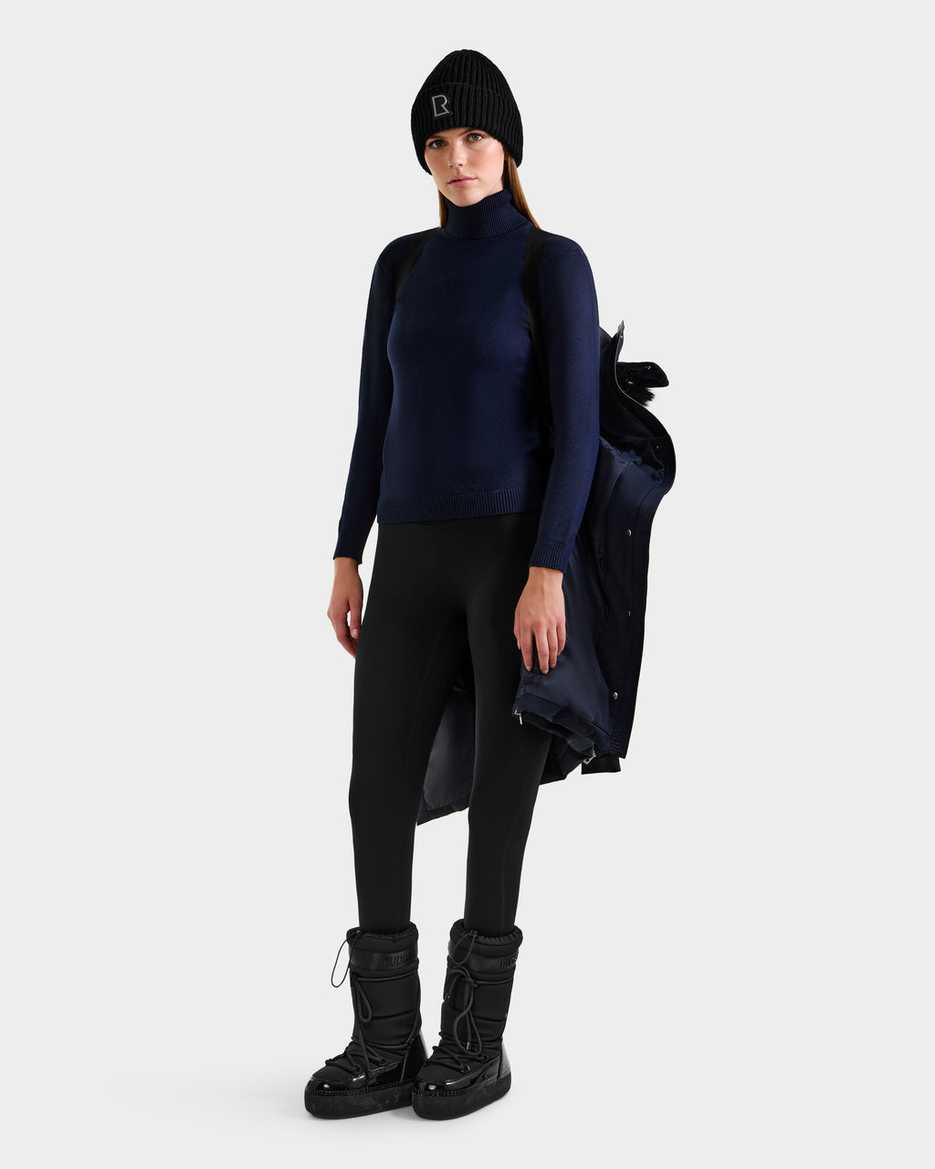 A woman holds the Rudsak KEHLANI H - 8125643 Navy RDS-certified down winter coat over one shoulder, dressed in a black beanie, navy turtleneck, black leggings, and black winter boots against a plain white background.