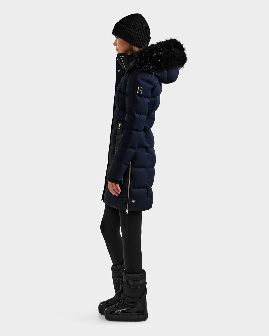 A woman in a dark winter coat stands in profile against a white background, highlighting the warmth of the Rudsak KEHLANI H - 8125643 Navy women's down parka.