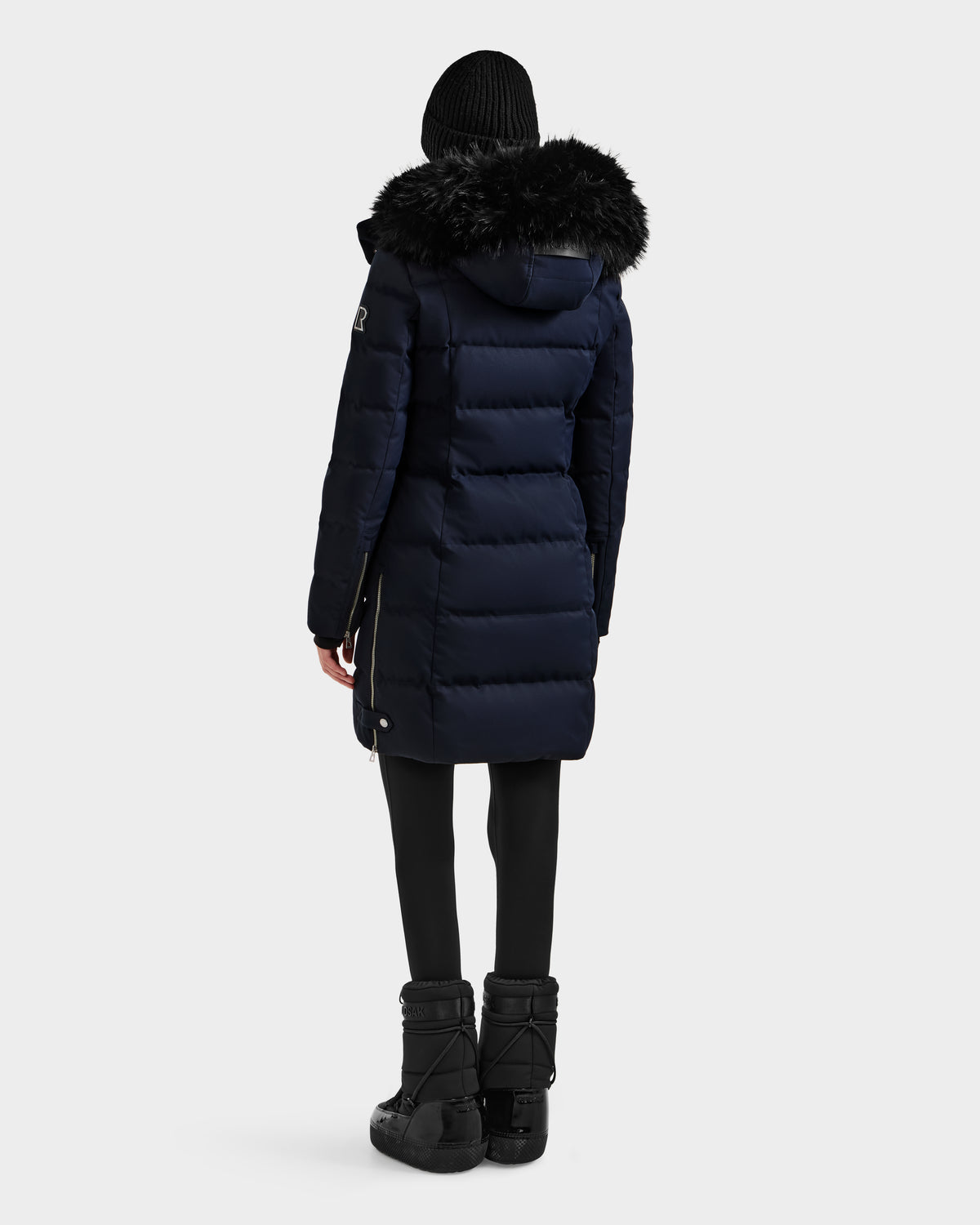 A person is seen from the back against a plain light background wearing Rudsak’s KEHLANI H - 8125643 Navy women’s down parka with RDS-certified down, a black fur-trimmed hood, black leggings, boots, and a black knit beanie.