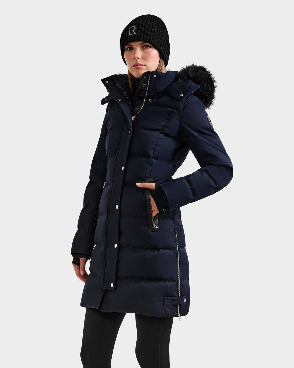 A woman wears the Rudsak KEHLANI H - 8125643 Navy down-filled parka with a fur-trimmed hood and a black beanie, standing forward with one hand in her pocket against a plain light background.