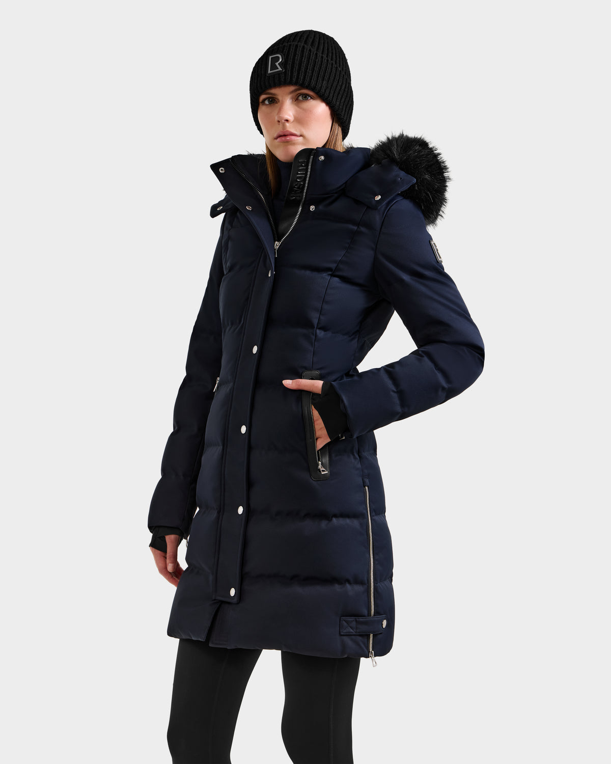 A woman wears the Rudsak KEHLANI H - 8125643 Navy down-filled parka with a fur-trimmed hood and a black beanie, standing forward with one hand in her pocket against a plain light background.